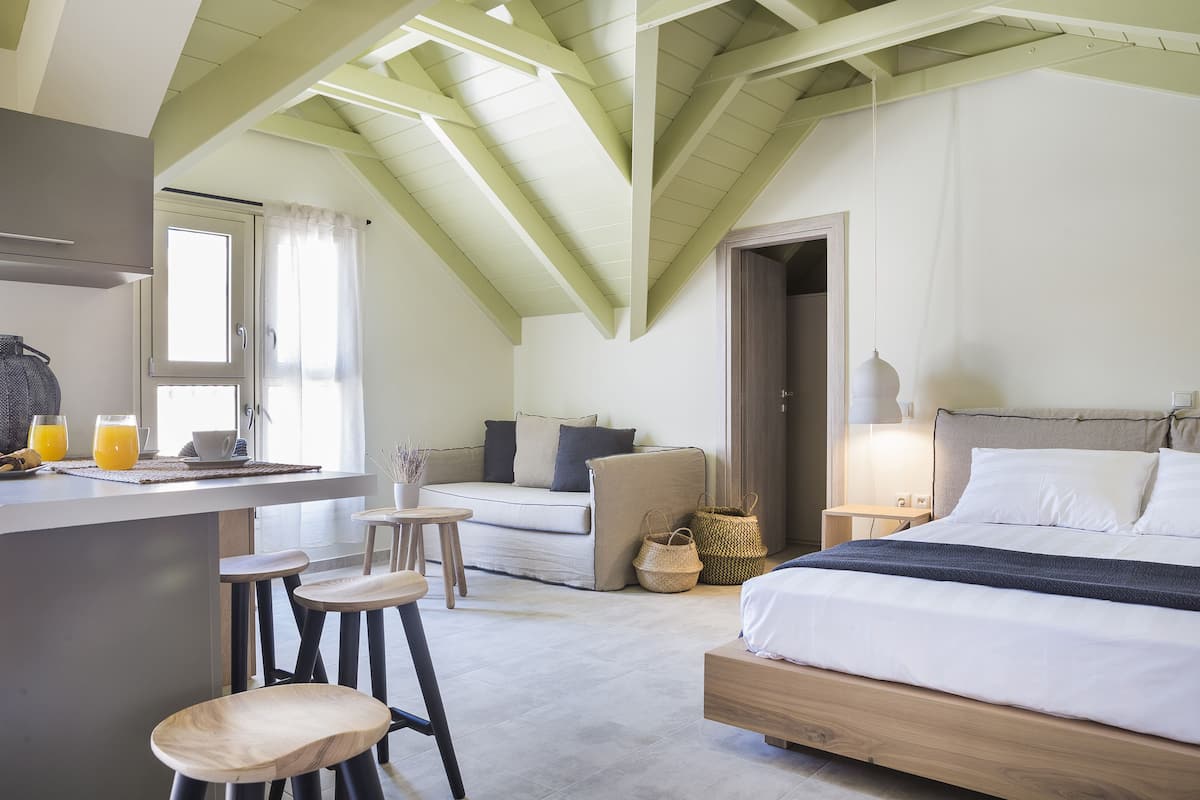 A modern bedroom with a loft-style ceiling, a large bed, a small seating area, and a kitchenette.