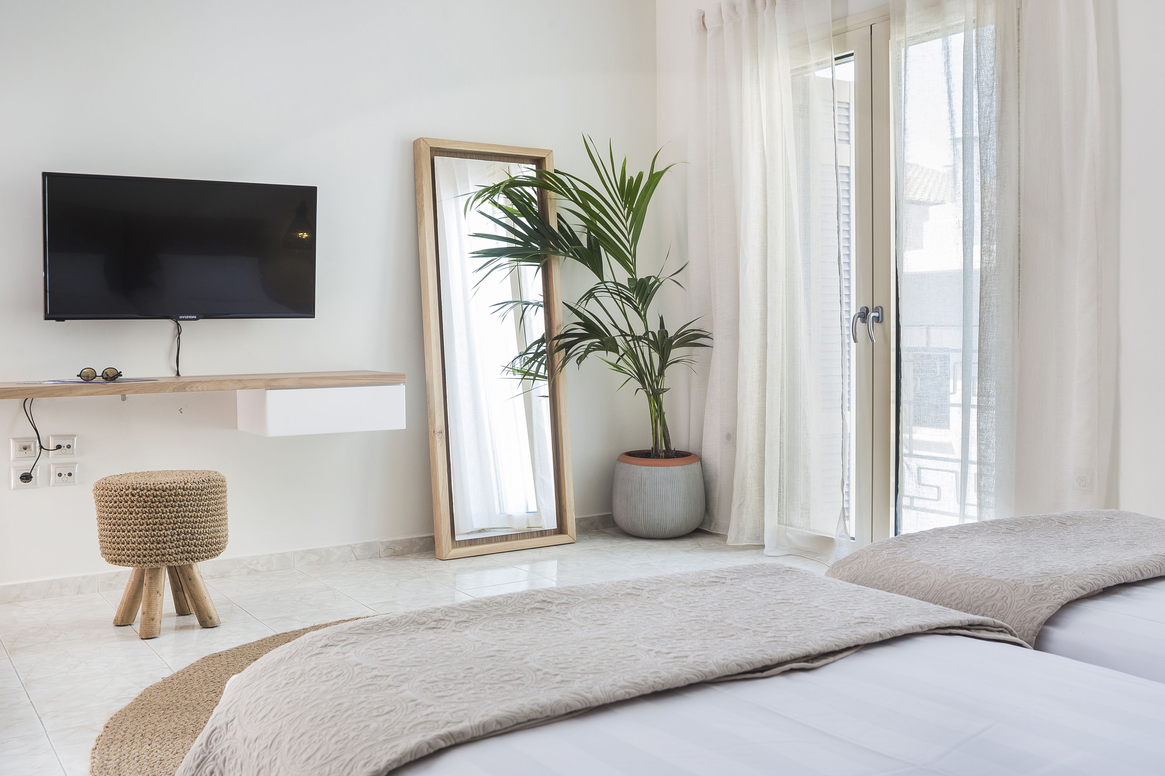 A modern hotel room with a flat-screen TV mounted on the wall, a large mirror, a potted plant, a small woven stool, and a bed with a beige blanket.