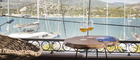 A wooden table with two glasses of a yellow beverage, a woven chair, and a view of boats and hills through a window.