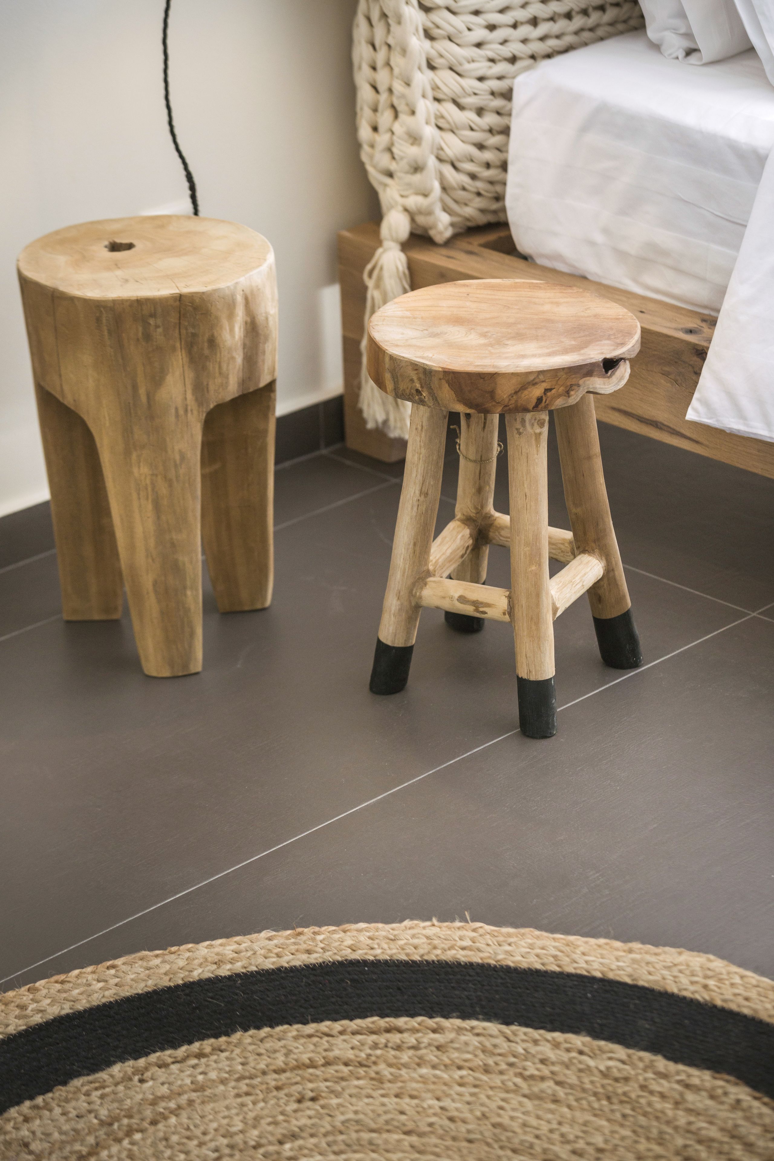 A wooden stool and a matching stool with a black base, placed on a dark tiled floor in a room with a woven rug.