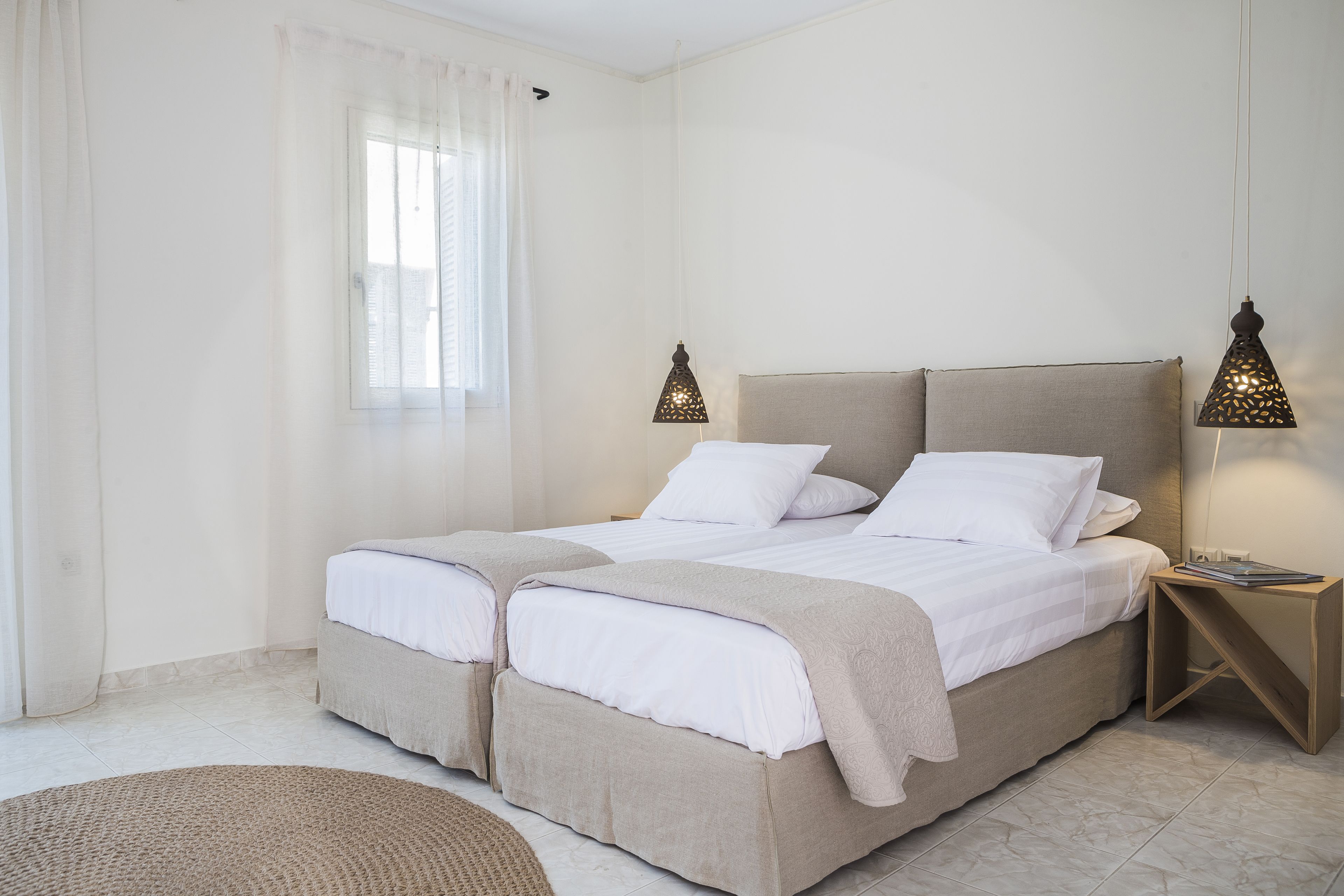 Una cama doble con cabecera, dos mesitas de noche, una ventana con cortinas traslúcidas y una lámpara de pie.