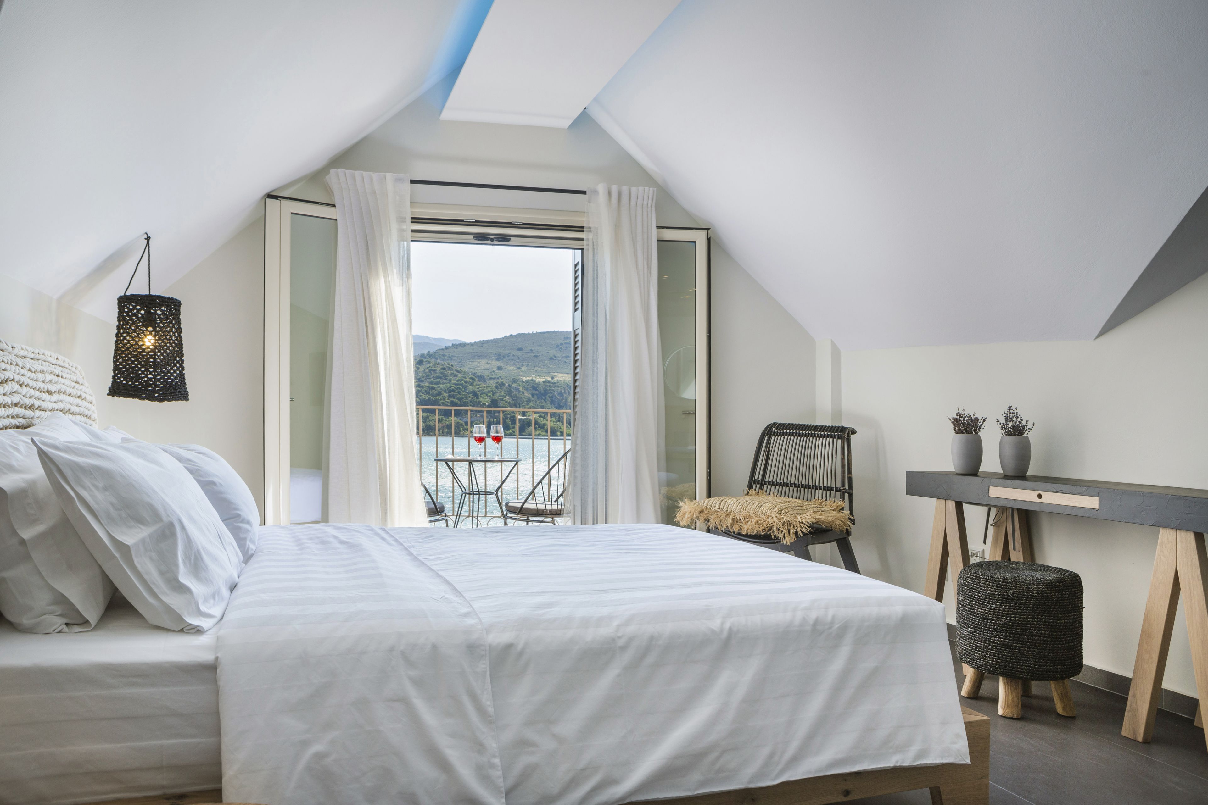 A bedroom with a bed, a desk, a chair, and a view of a balcony with a table and chairs.