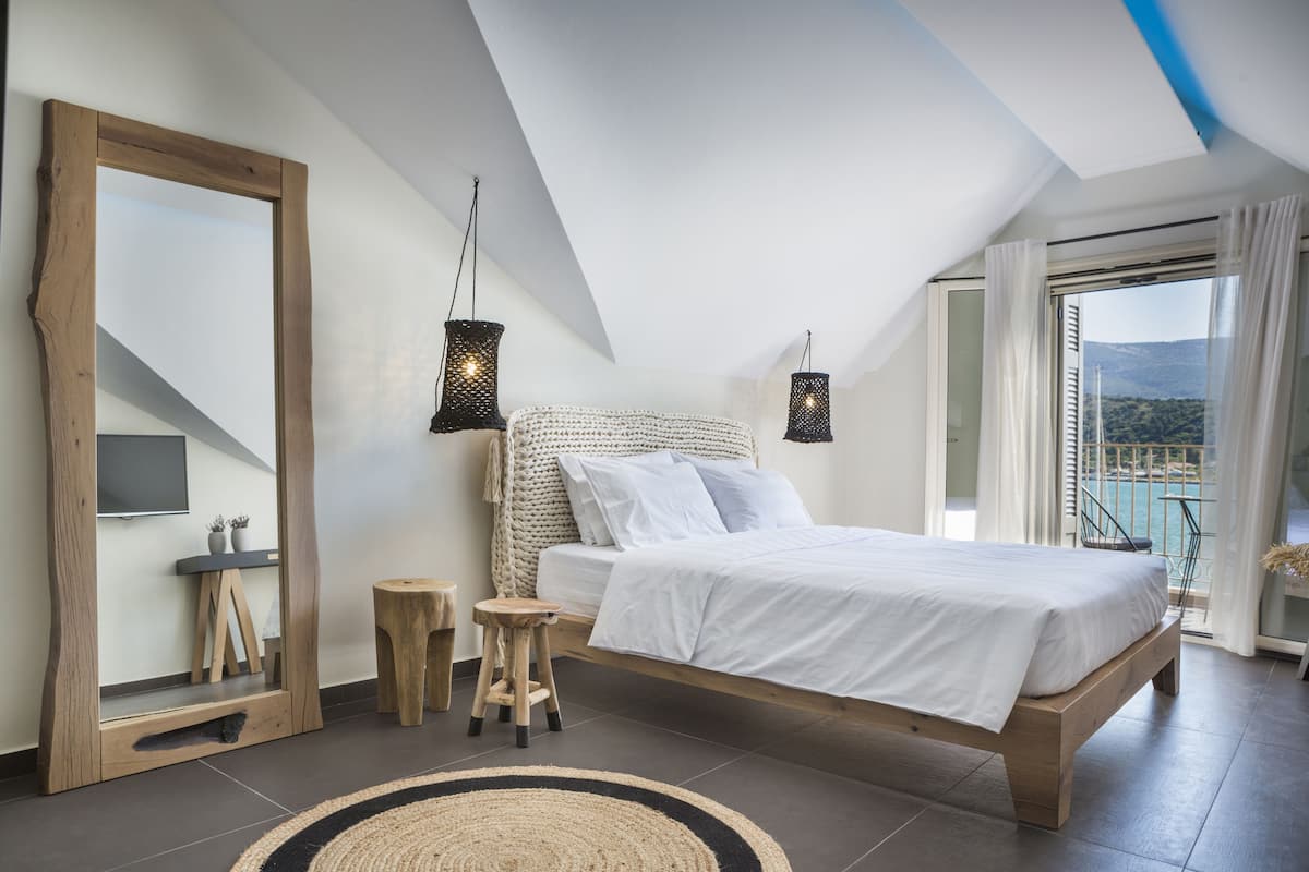 A bedroom with a large bed, a wooden-framed mirror, two hanging lights, a small stool, and a view of the outdoors through a sliding door.