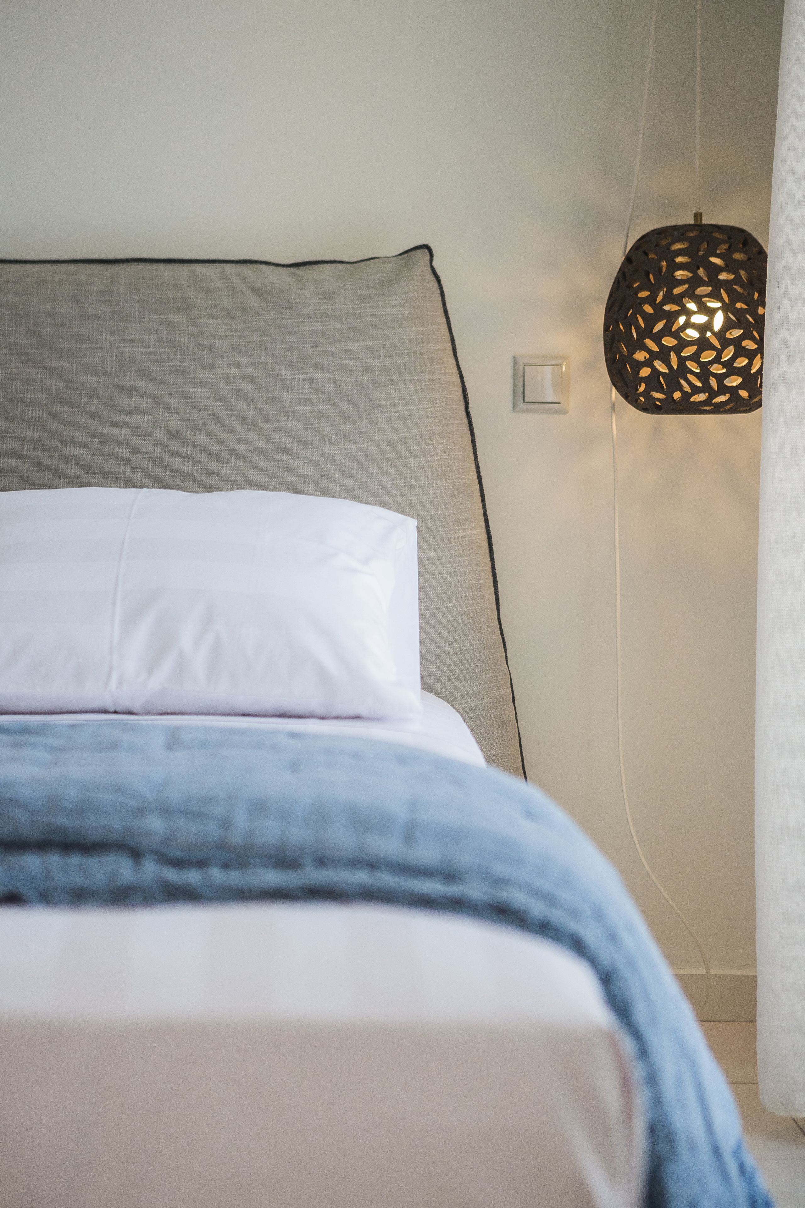 A neatly made bed with white bedding and a blue throw blanket, a textured grey headboard, and a wall-mounted lamp with a patterned shade.