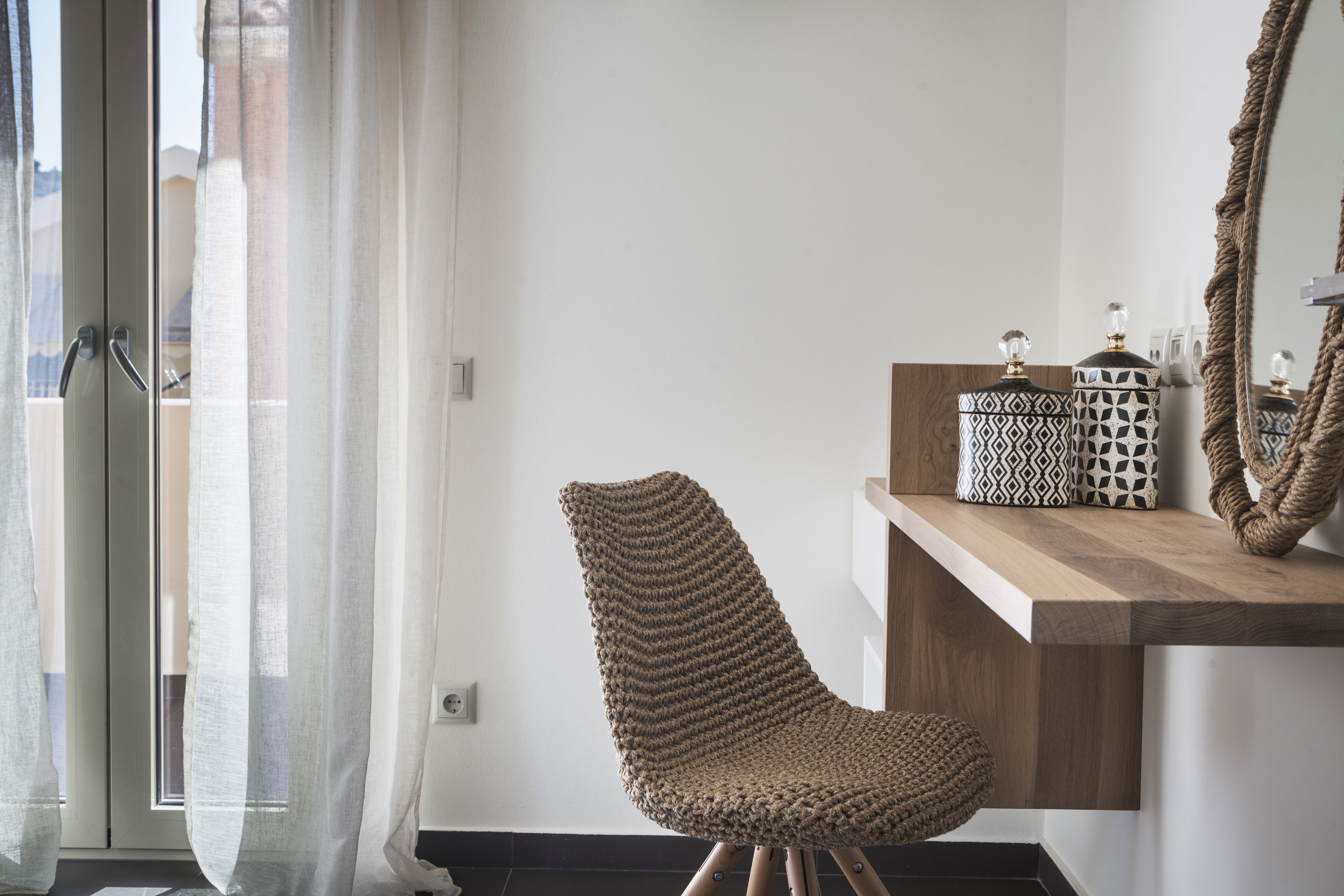 Een moderne kamer met een groot raam, een geweven stoel, een houten bijzettafeltje met decoratieve objecten en een spiegel.