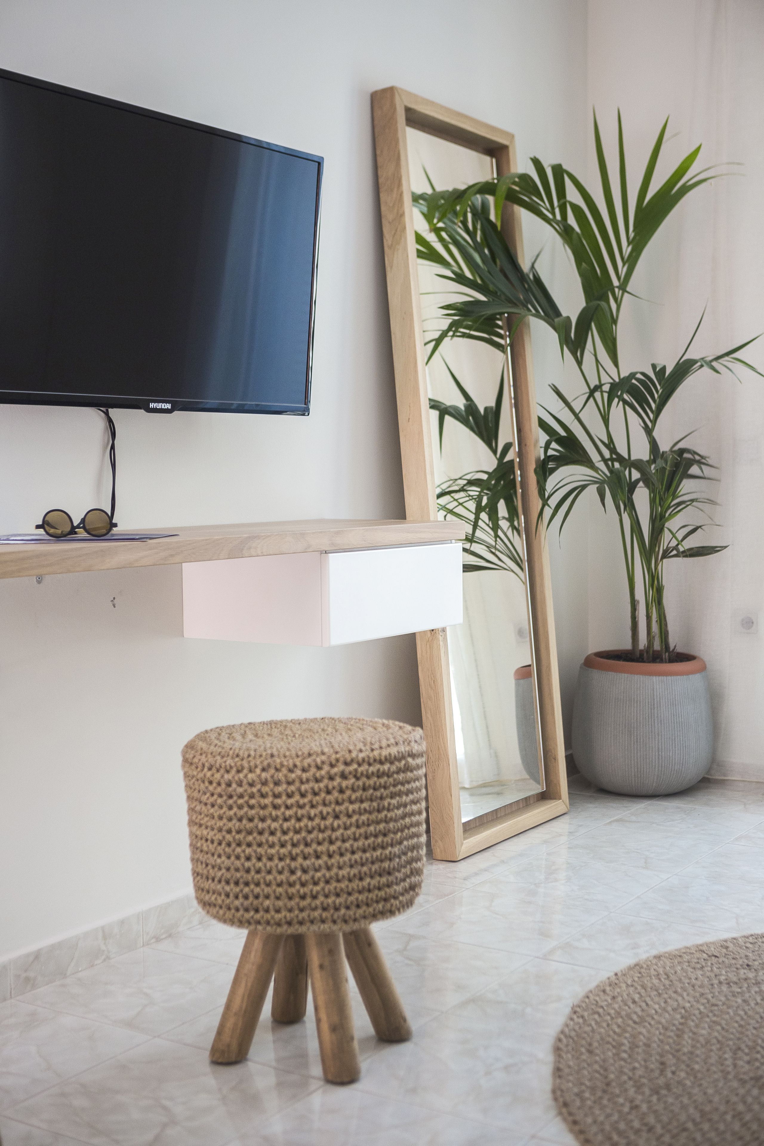 A modern living room with a flat-screen TV, a wooden TV stand, a large mirror, a potted plant, and a woven stool.