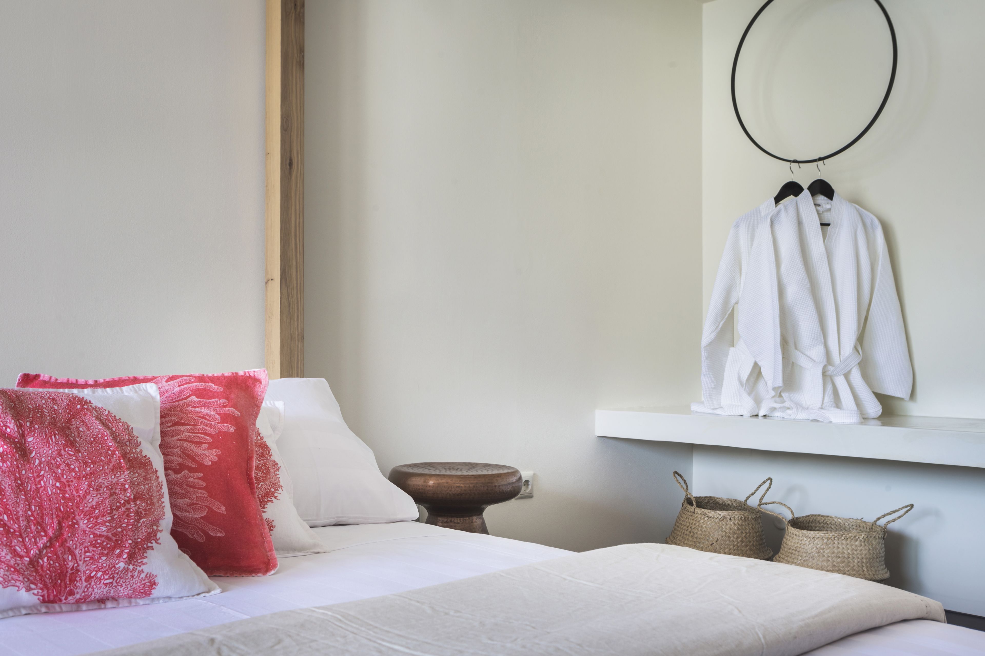 A neatly made bed with white bedding, a wooden nightstand with a bowl, and a shelf with white robes and baskets.
