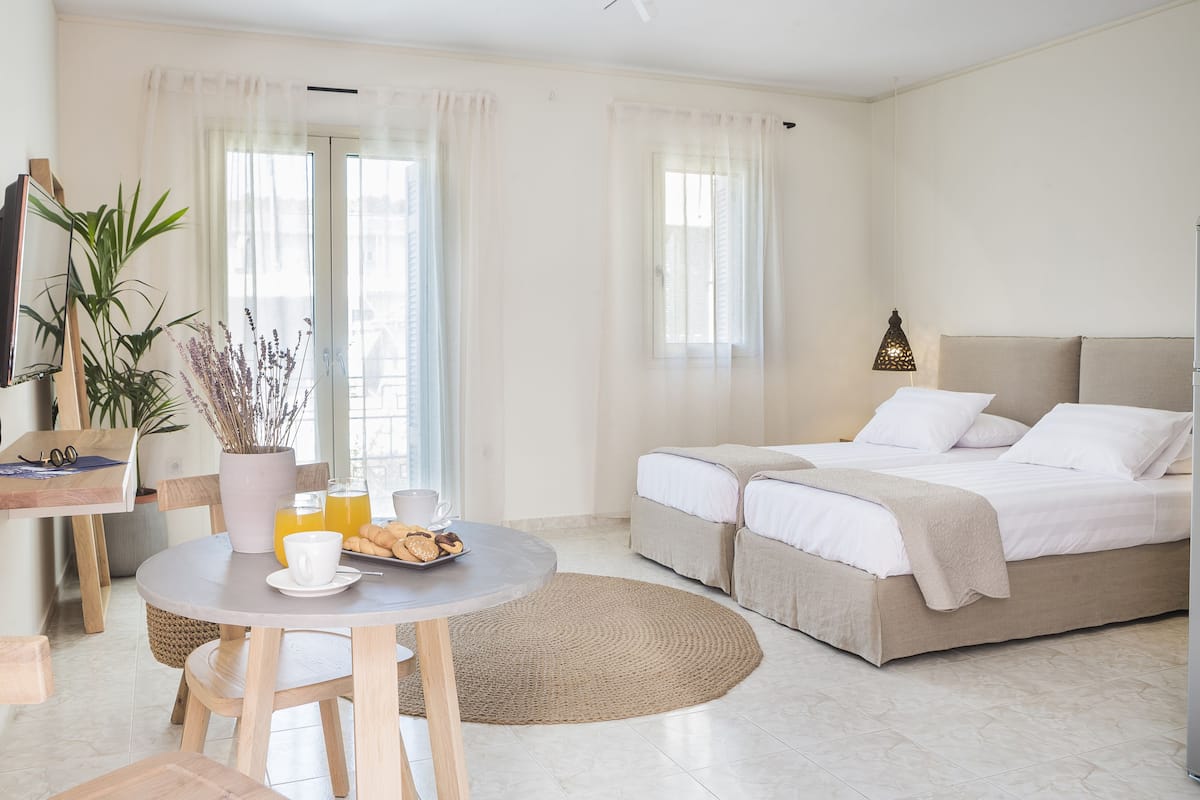 A modern hotel room with a bed, a dining area with a table and chairs, and a large window with curtains.