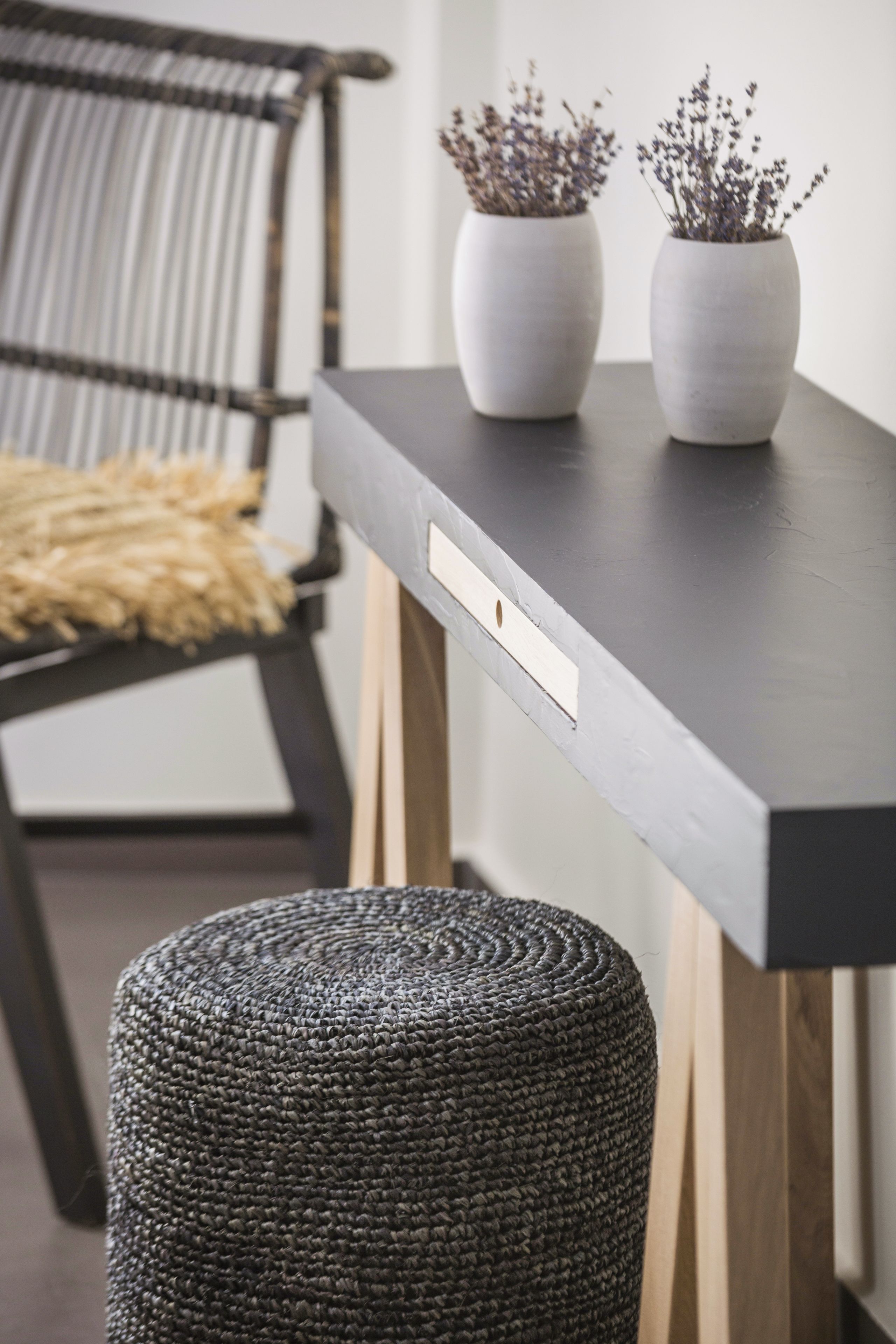A modern room with a dark wooden table, two potted plants, a woven ottoman, and a wicker chair with a cushion.