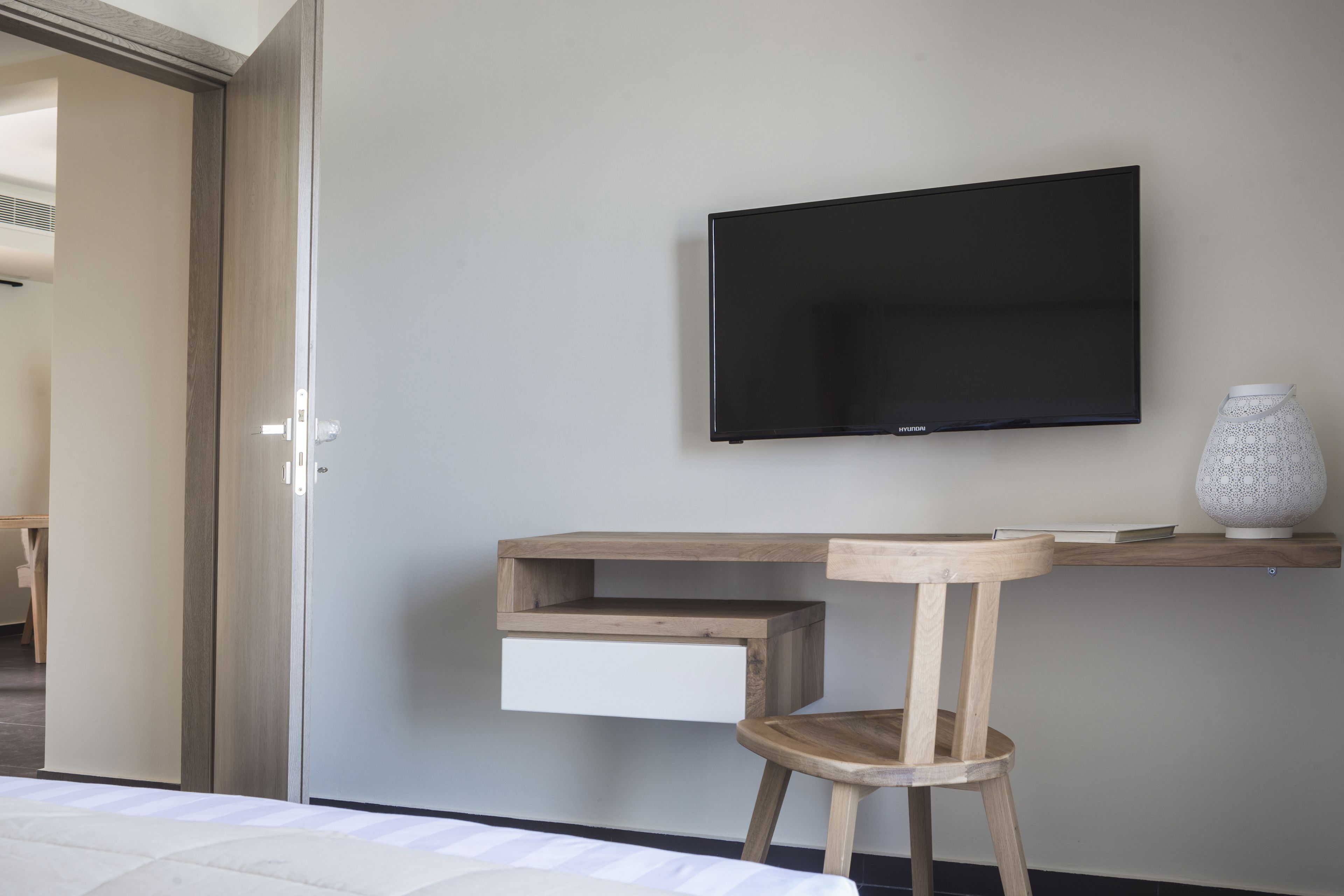 A bedroom with a bed, a wooden desk with a TV, a chair, and a small shelf with a vase.