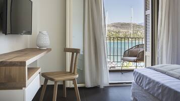 A room with a wooden desk, a chair, a bed, and a balcony with a view of a body of water and a hill.