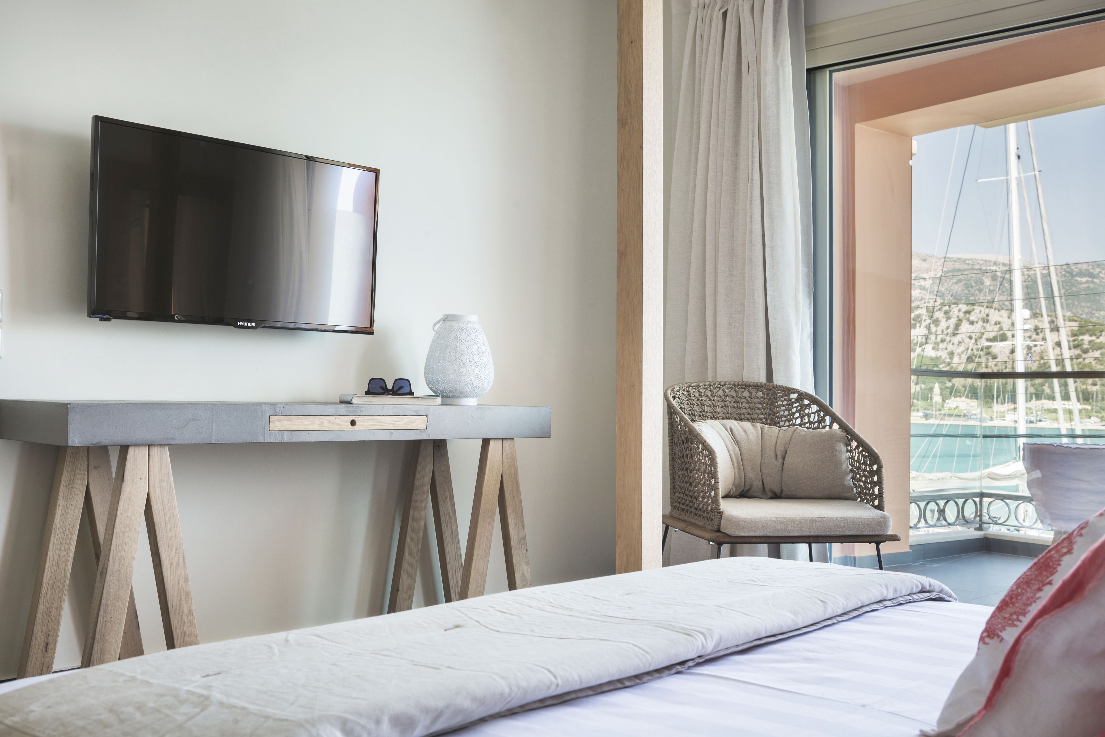 A hotel room with a bed, a flat-screen TV, a small table with a vase, a wicker chair, and a balcony with a view of the sea.