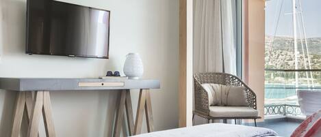 A hotel room with a bed, a flat-screen TV, a small table with a vase, a wicker chair, and a balcony with a view of the sea.