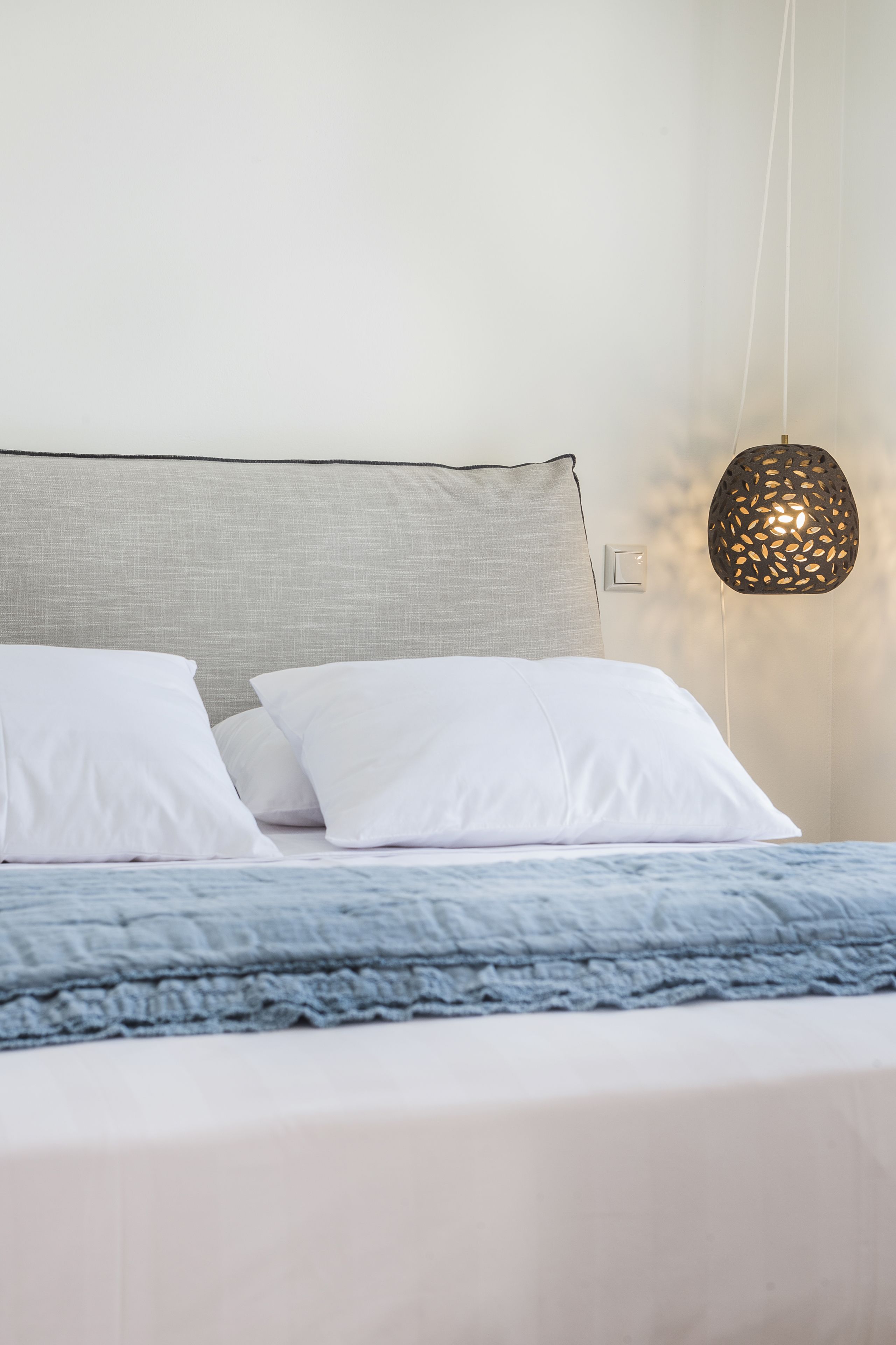Un lit bien fait, avec des oreillers blancs et une couverture bleue, une tête de lit grise et un luminaire suspendu.