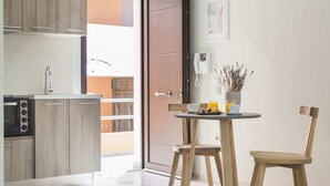 A modern kitchen with wooden cabinets, a microwave, and a sink. There is a dining area with a table, two chairs, and a vase with dried plants. A door leads to an outdoor balcony. - Argostoli Marina Suites (Kefalonia)