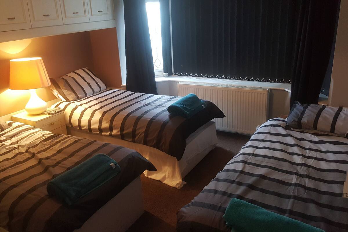 A hotel room with two beds, a nightstand with a lamp, a window with curtains, and a radiator.