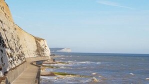 A coastal walkway with a stone wall and a wooden deck leading to the sea. - John Wayne Lodge (Peacehaven)
