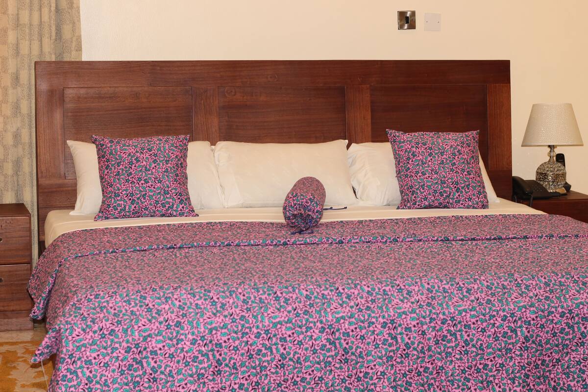A bed with a floral-patterned bedspread, two pillows, a nightstand with a lamp, and a wooden headboard.