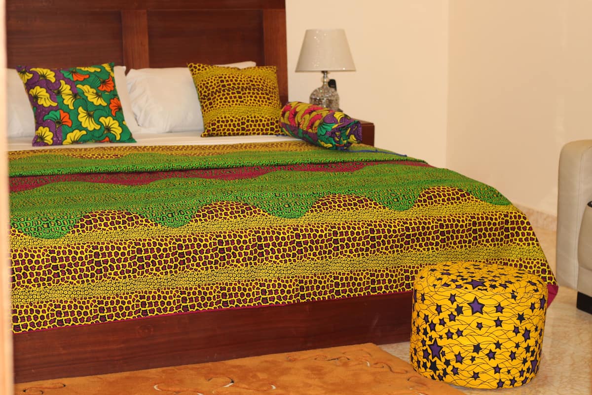 A bed with a colorful, patterned bedspread and pillows, a bedside lamp, and a small ottoman with a star pattern.