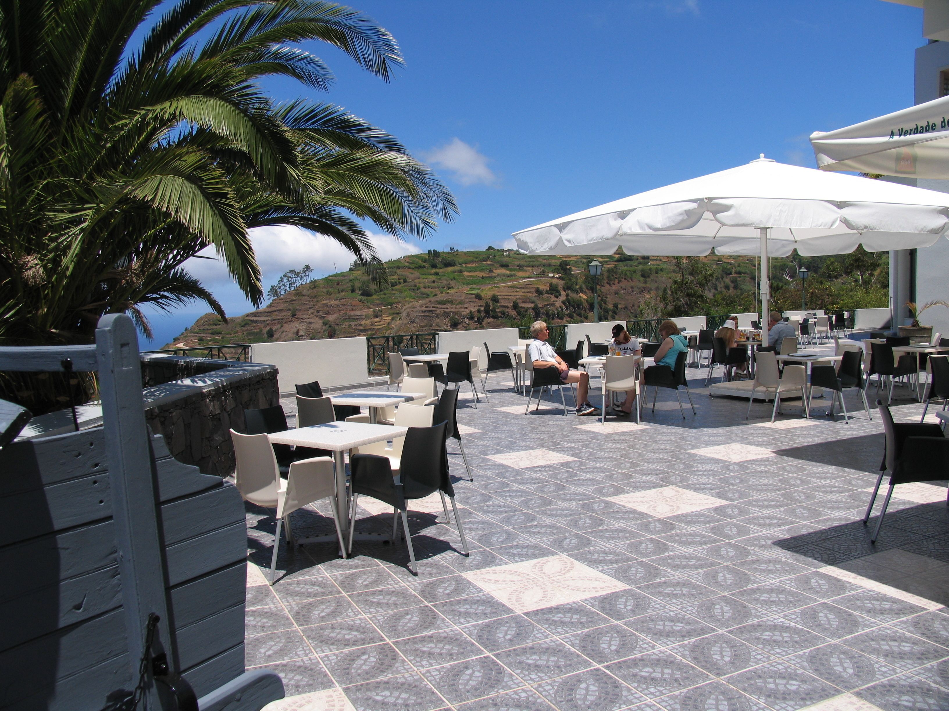 An outdoor patio with tables and chairs, a white umbrella, and a view of the hillside.