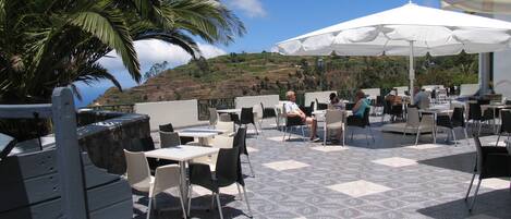 An outdoor patio with tables and chairs, a white umbrella, and a view of the hillside.