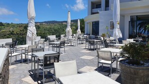 An outdoor patio with tables and chairs, white umbrellas, and a view of the hills. - Hotel Jardim Atlantico (Calheta)