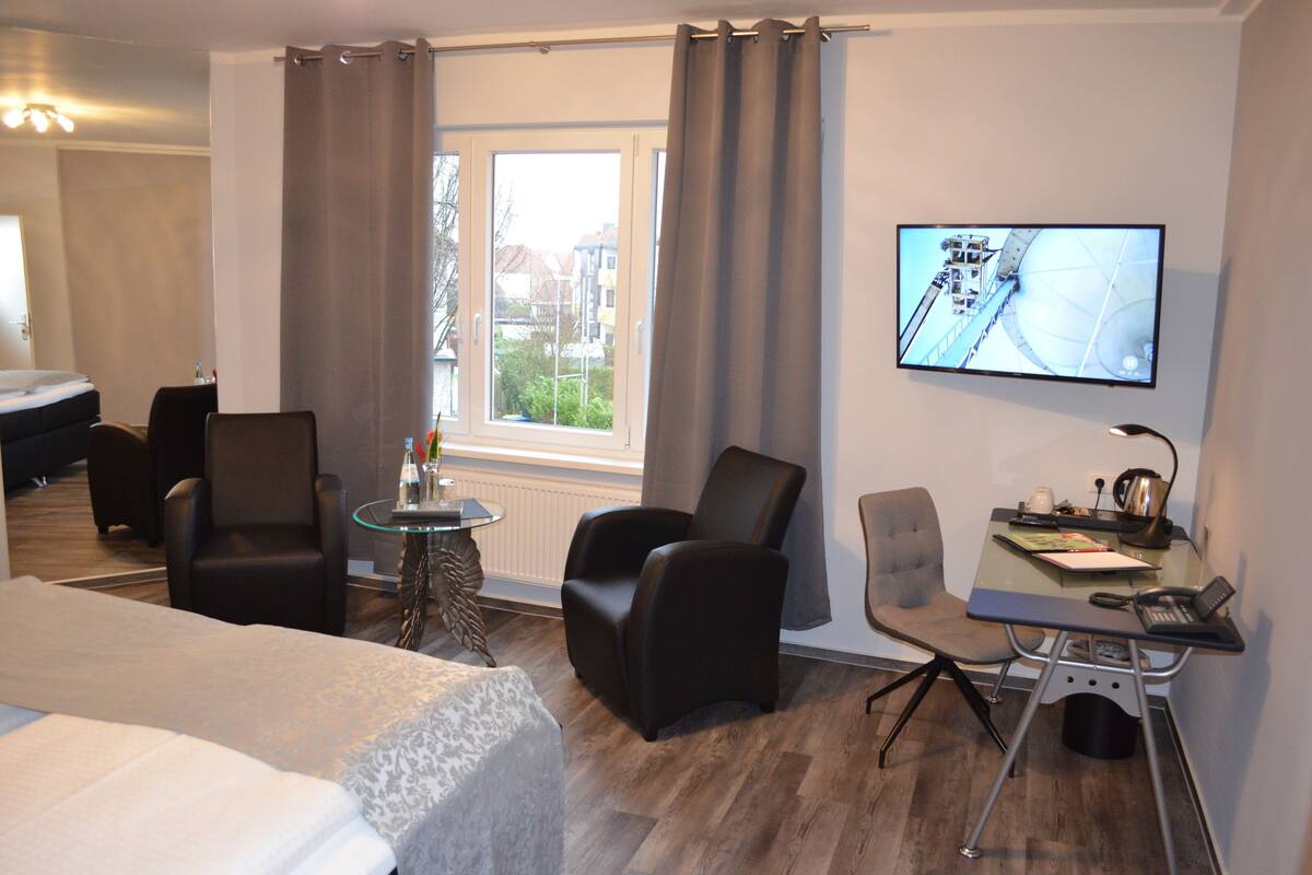 Une chambre d’hôtel comprenant un lit, deux fauteuils, une petite table, un bureau avec une chaise, une télévision et une fenêtre avec des rideaux.