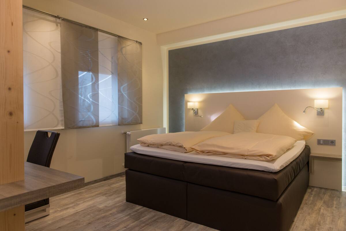 Une chambre moderne avec un grand lit, des lampes de chevet et une vue sur la ville, visible à travers une fenêtre ornée d’un rideau.
