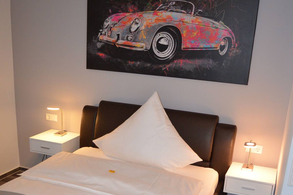 Un lit bien fait, avec un oreiller, une table de chevet sur laquelle se trouve une lampe, et un tableau représentant une voiture accroché au mur.