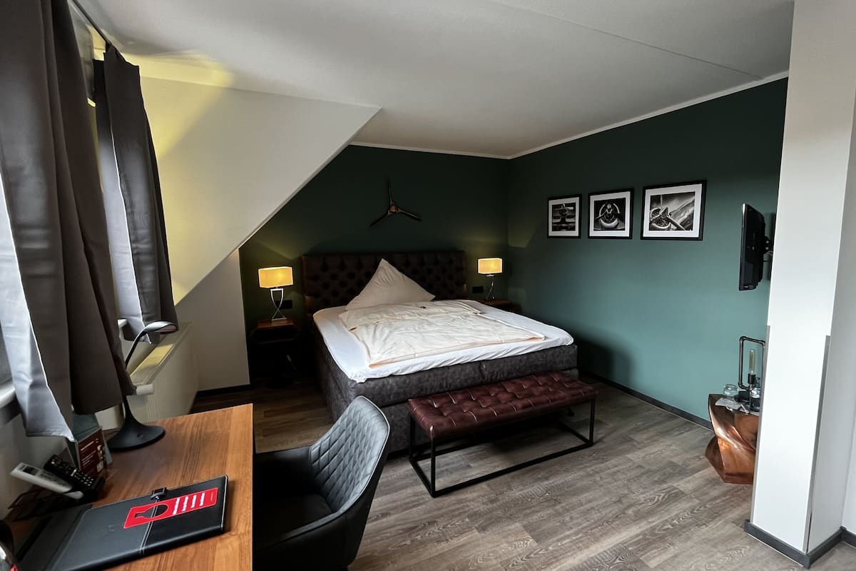 Une chambre d’hôtel avec un lit, un bureau avec un ordinateur, une chaise, un banc, une télévision et des tableaux encadrés au mur.