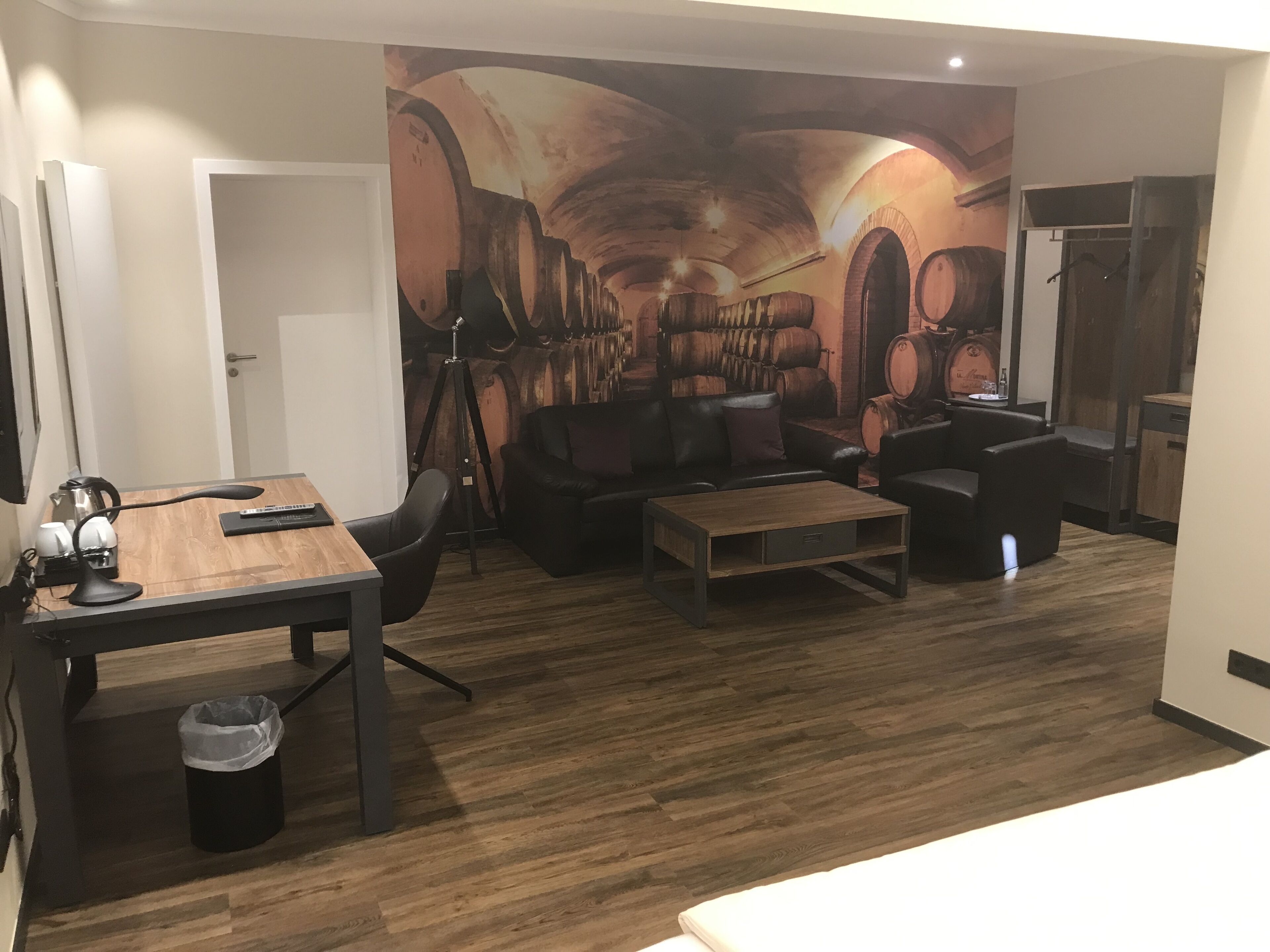 Ein modernes Wohnzimmer mit Holzboden, einer schwarzen Ledersofa, einem Holztisch und einem Esstisch mit Stuhl. An der Wand befindet sich ein großes Wandbild, das Fässer und einen Tunnel zeigt.