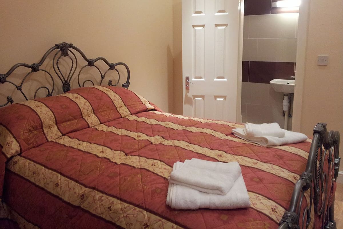 Uma cama com colcha vermelha e bege, cabeceira de metal e uma porta branca ao fundo.