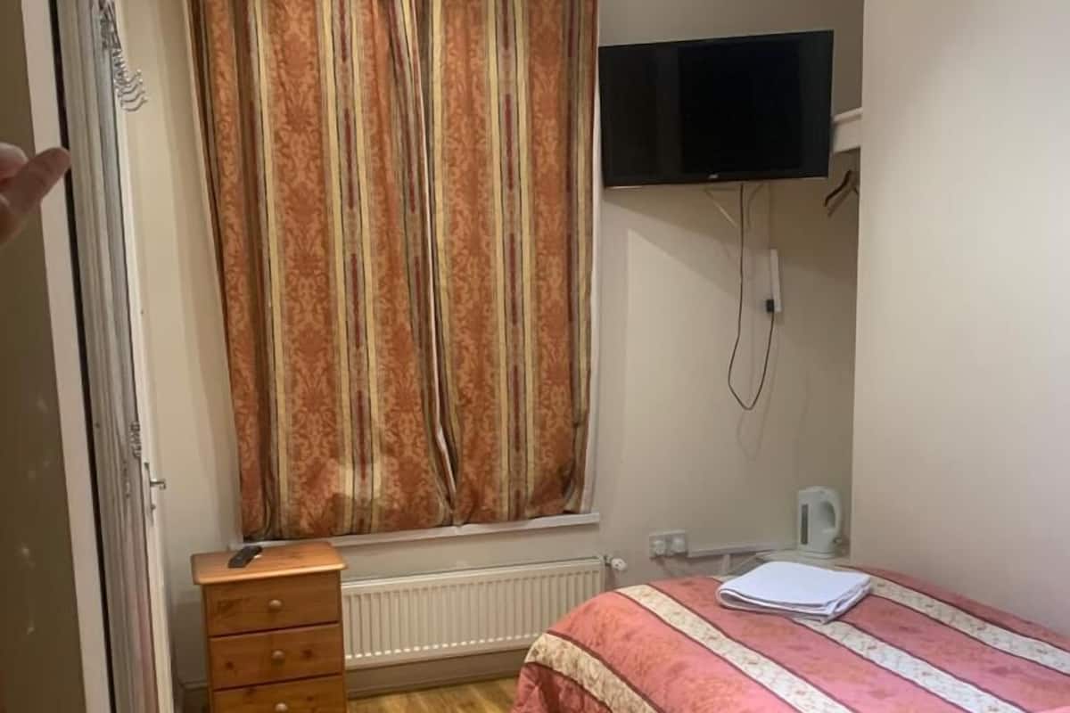 Quarto com piso de madeira, cama com colcha estampada, janela com cortinas, TV fixada na parede e radiador.