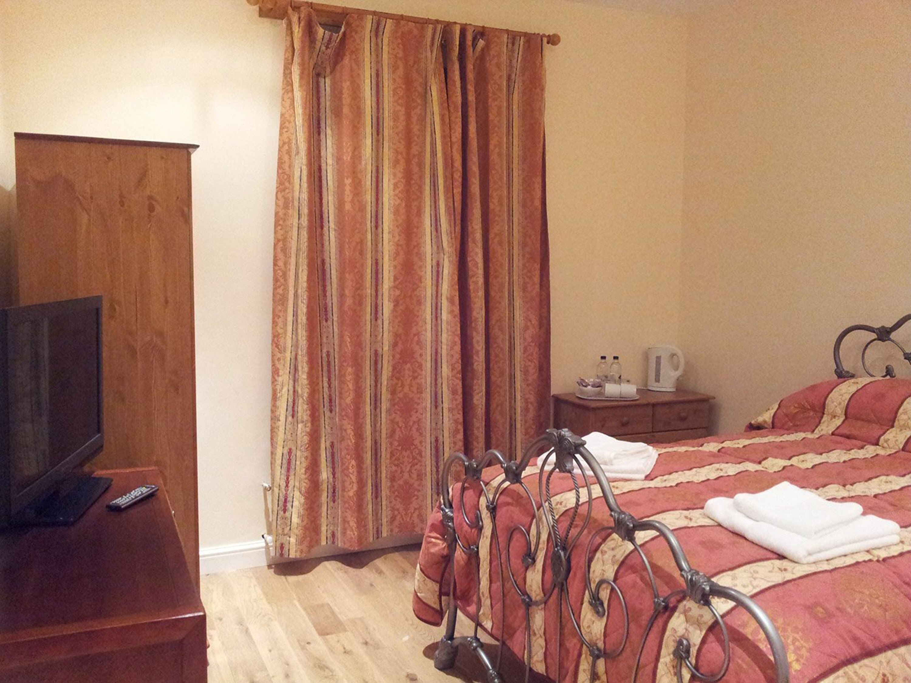 A bedroom with a wooden TV stand, a bed with a metal frame, a nightstand with a kettle, and a curtain with a striped pattern.