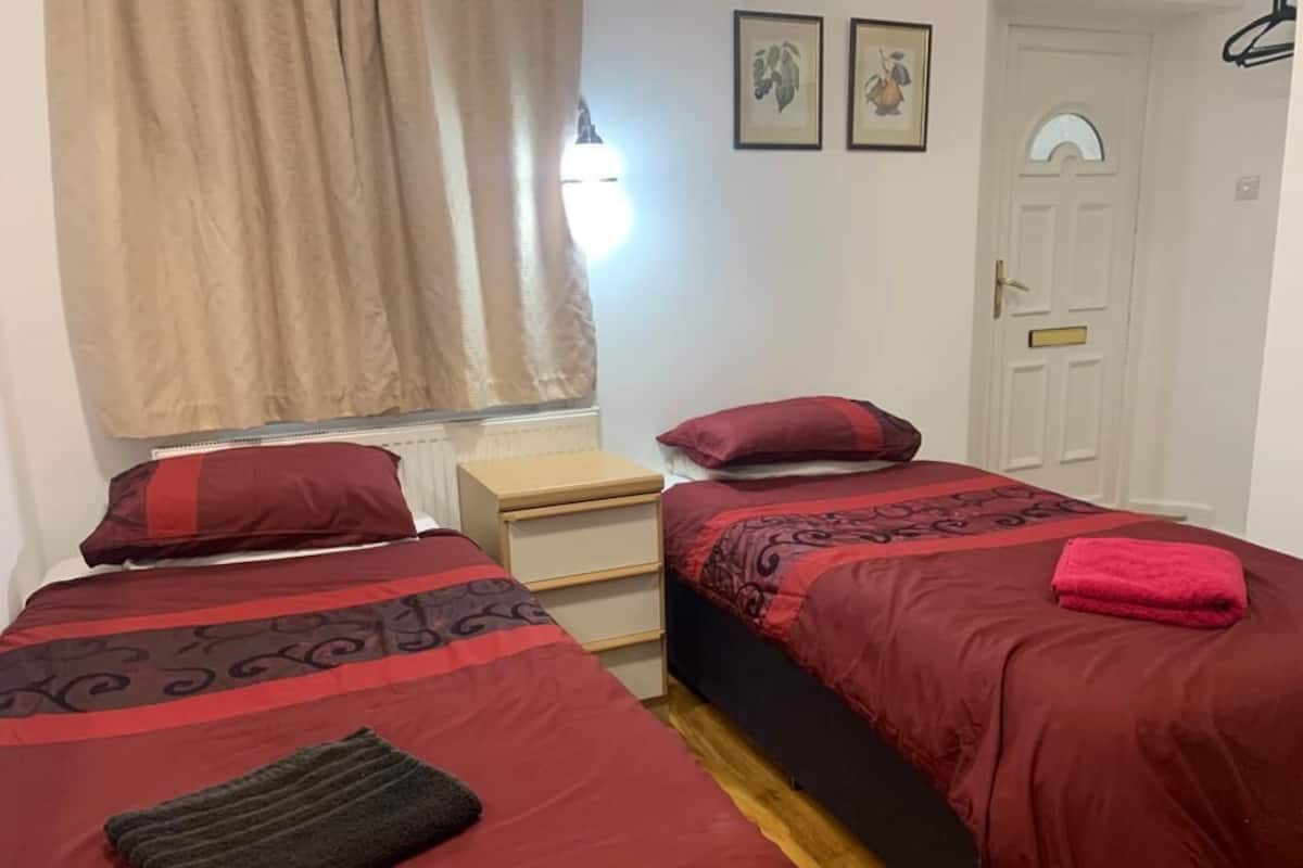 Quarto com duas camas, uma mesinha de cabeceira, um radiador, uma janela com cortinas e uma porta.