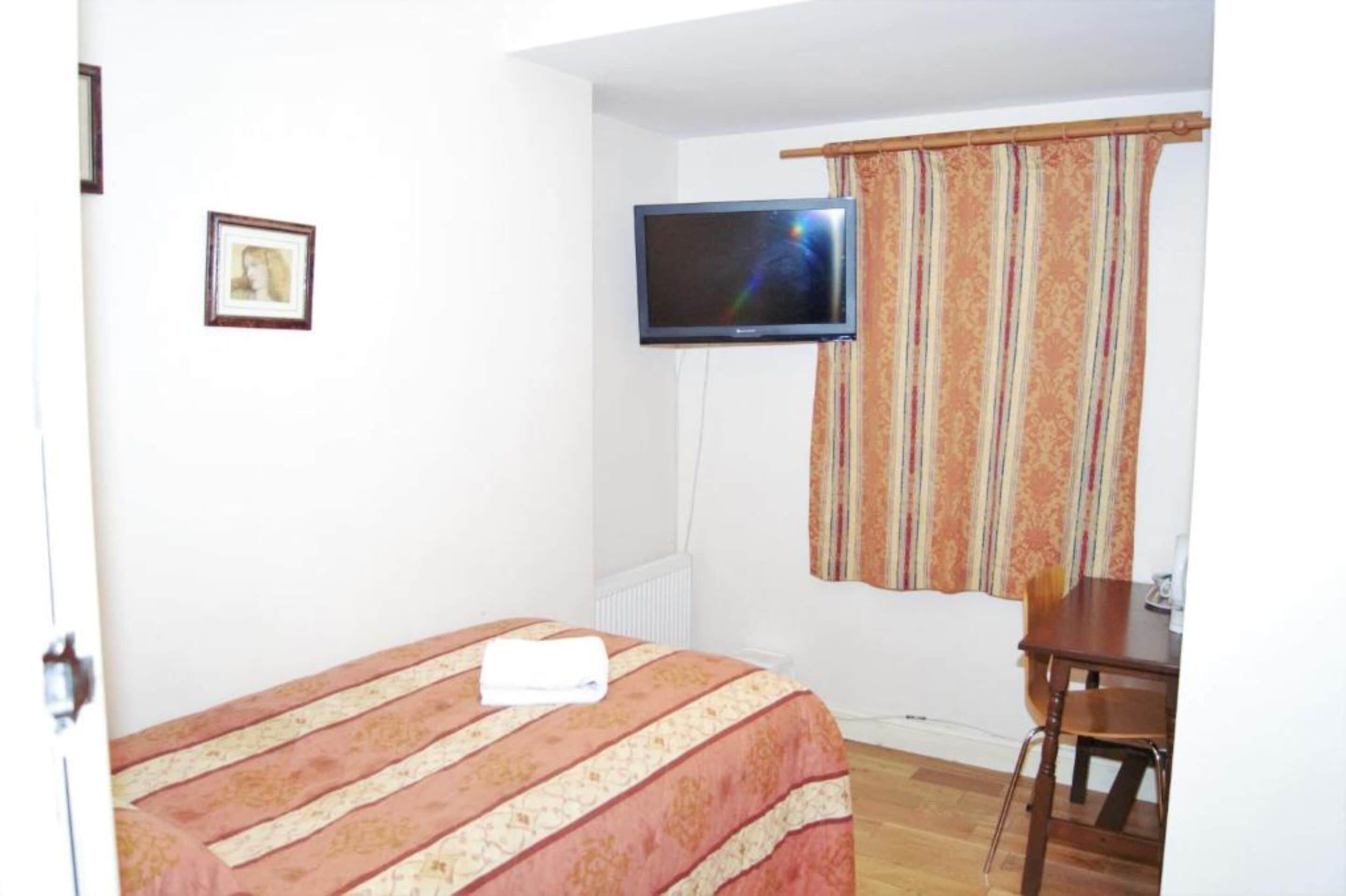 Quarto com cama, televisão na parede, janela com cortinas e cadeira de madeira.