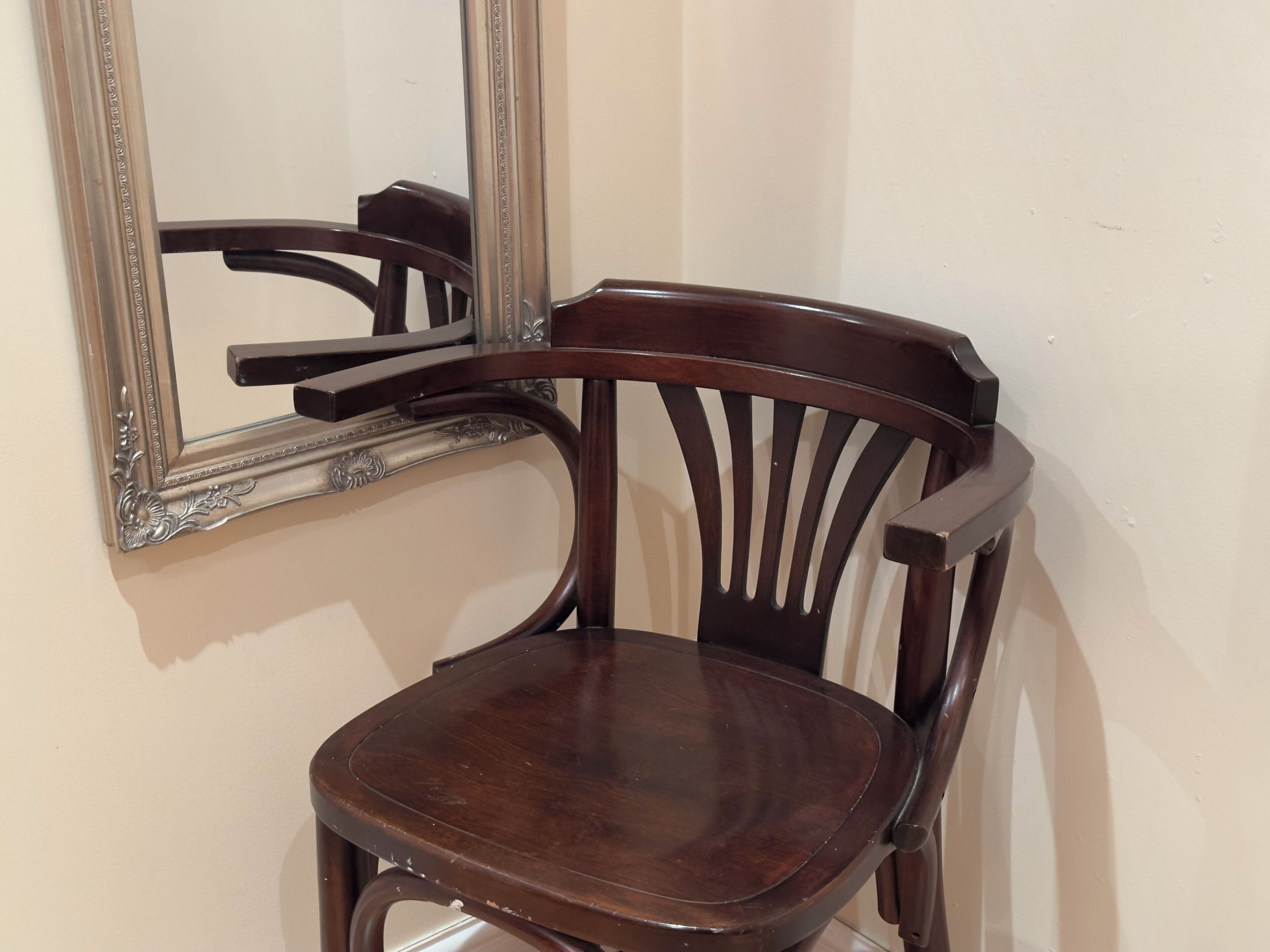 A wooden chair with a high backrest and a cushioned seat, positioned against a wall with a large mirror reflecting part of the room.