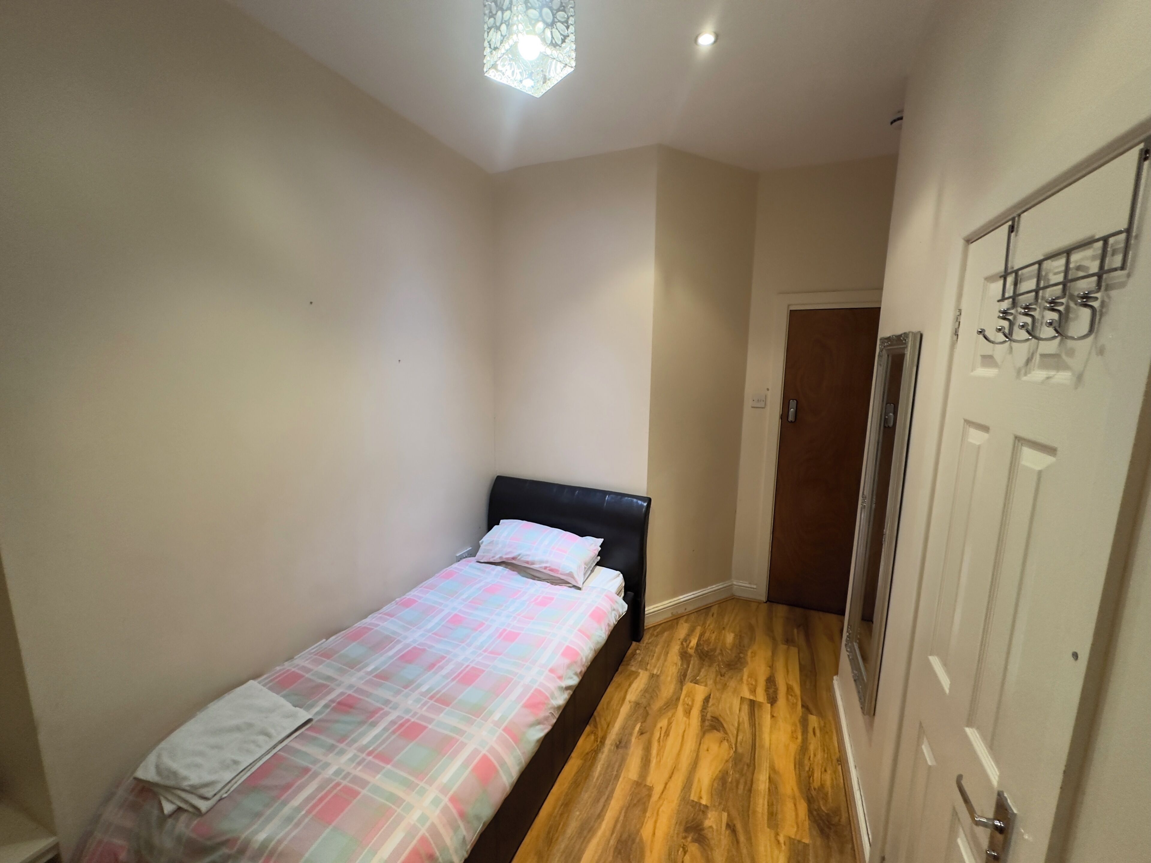 A small, single-bed room with a wooden floor, a plain wall, and a door with a coat rack.