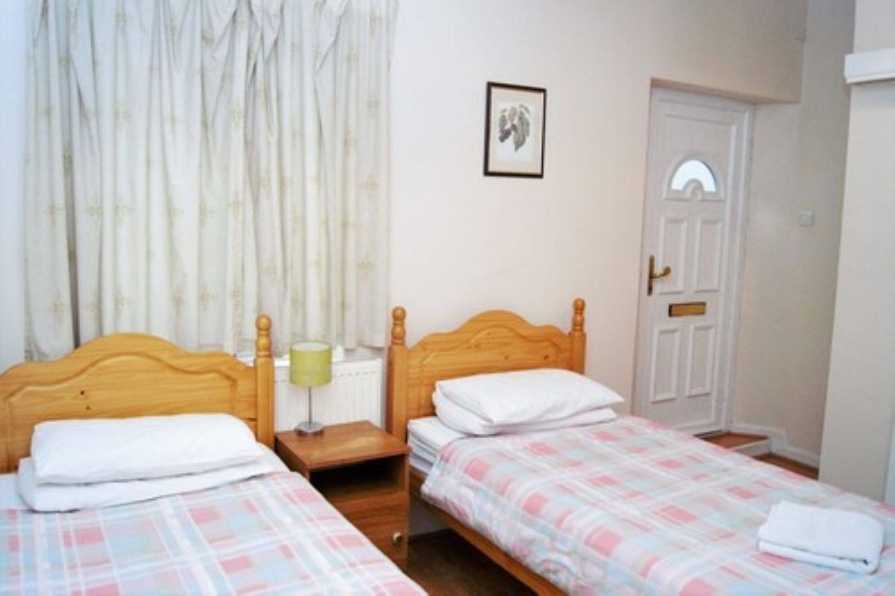 A bedroom with two single beds, a wooden headboard, a nightstand with a lamp, a framed picture on the wall, and a white door with a window.