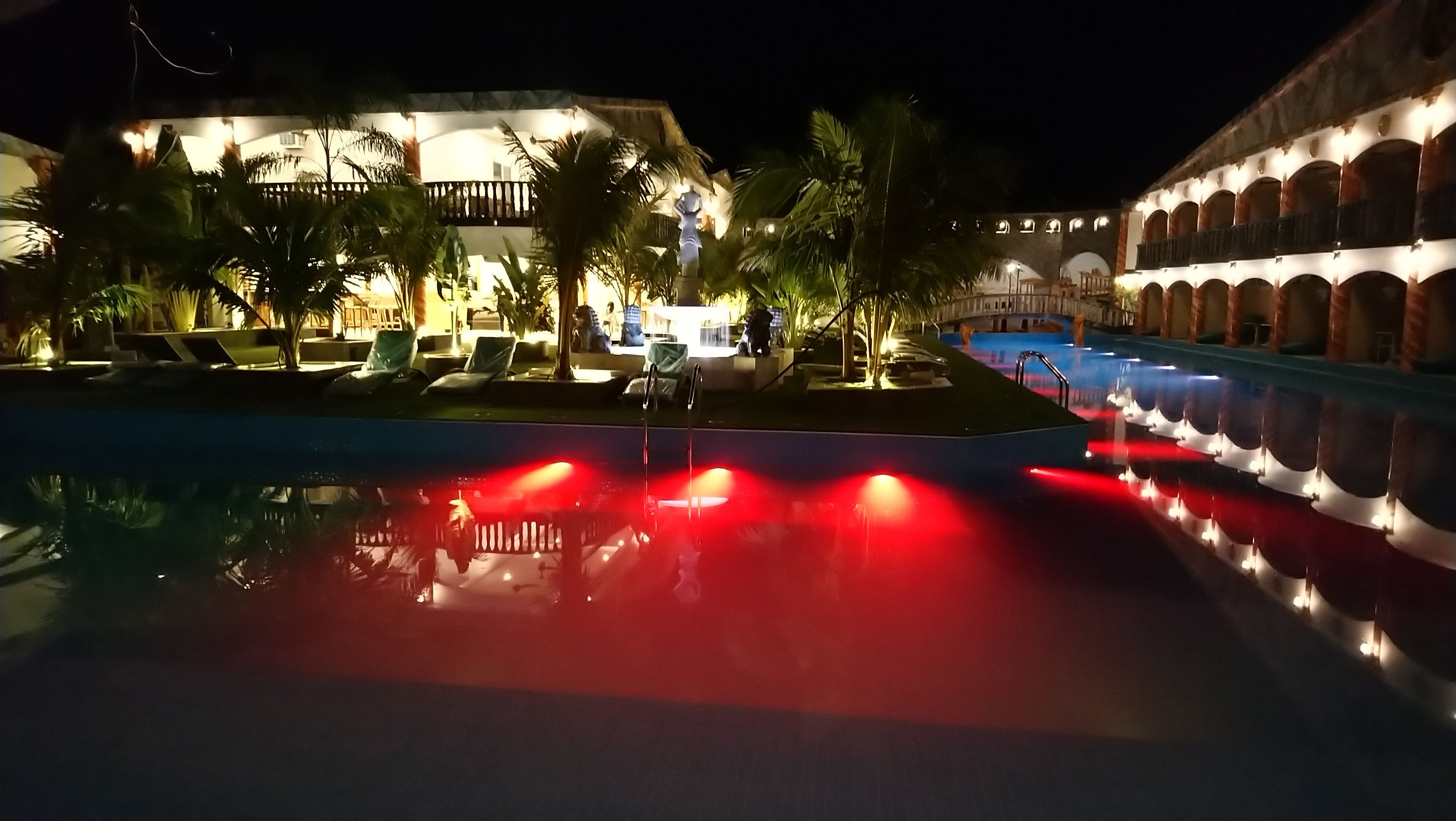 A nighttime pool area with a reflection of lights, palm trees, and a building with arches.