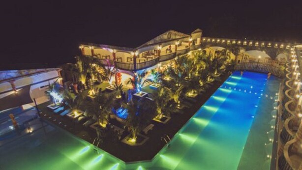 A large, illuminated swimming pool with a surrounding deck area, surrounded by palm trees and a building with multiple balconies.