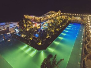 A large, illuminated swimming pool with a surrounding deck area, surrounded by palm trees and a building with multiple balconies.