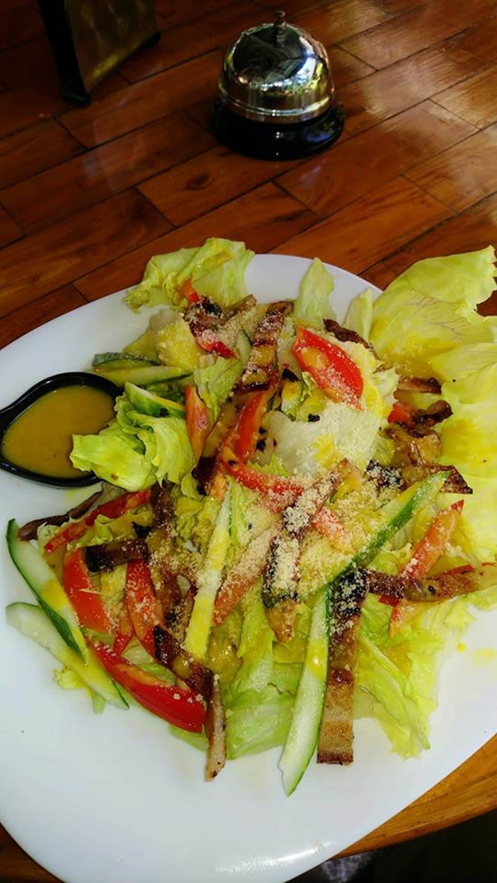 A plate of mixed greens salad with red peppers and croutons, accompanied by a small bowl of dressing, on a wooden table.