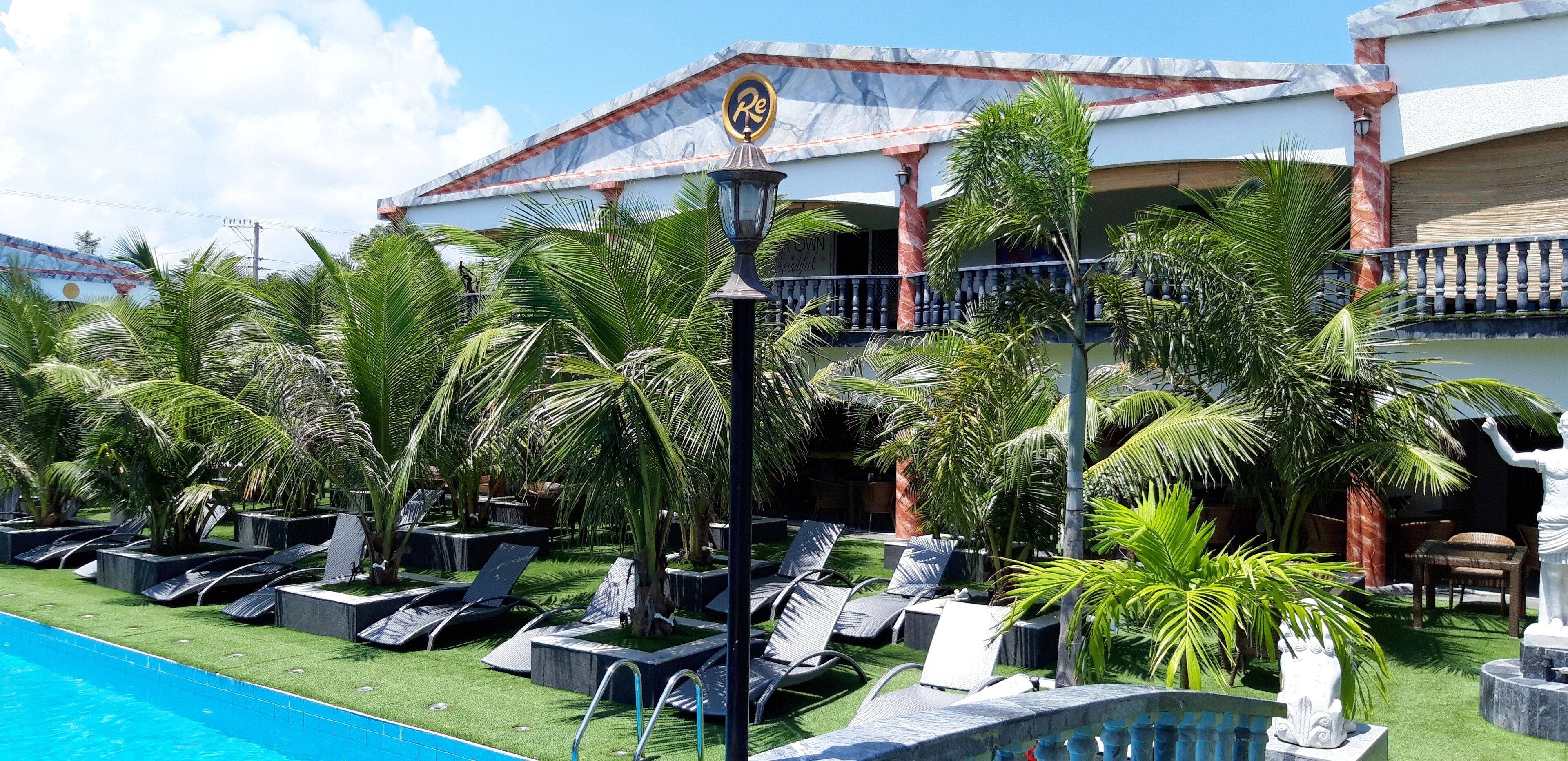 A poolside area with lounge chairs and palm trees, a statue, and a building with a balcony in the background.
