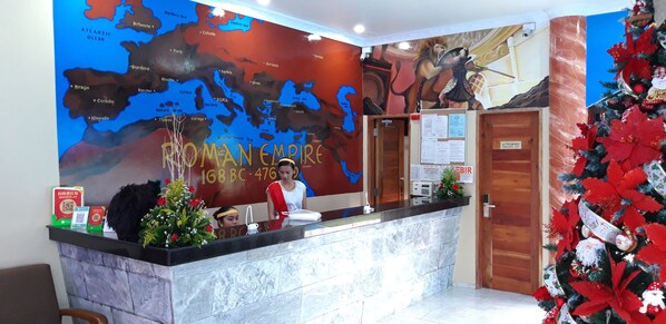 A hotel reception area with a large Roman Empire-themed wall map, a Christmas tree, and a receptionist.