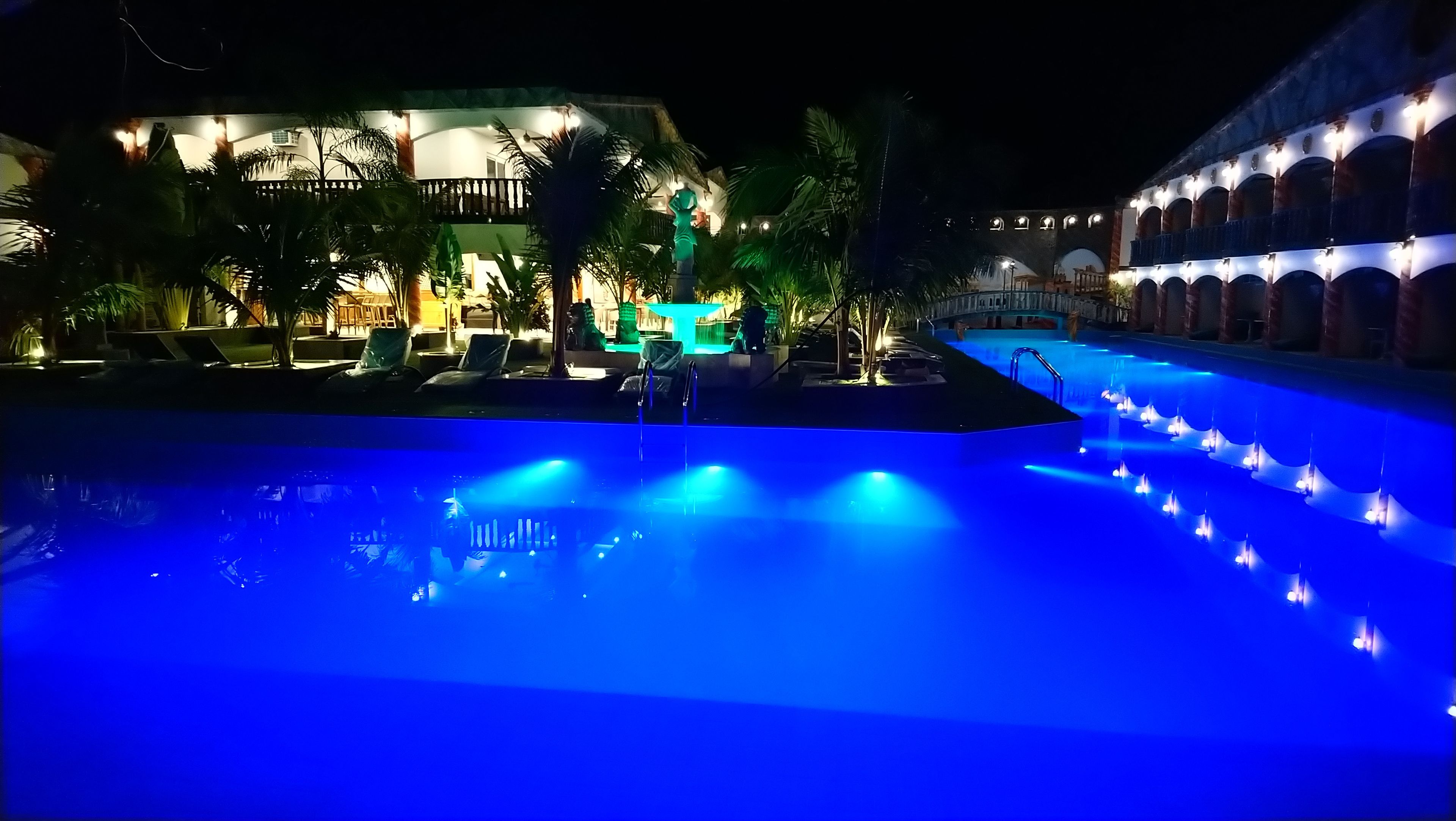 A nighttime pool area with a blue-lit water feature, lounge chairs, and a building with arched windows and a balcony.