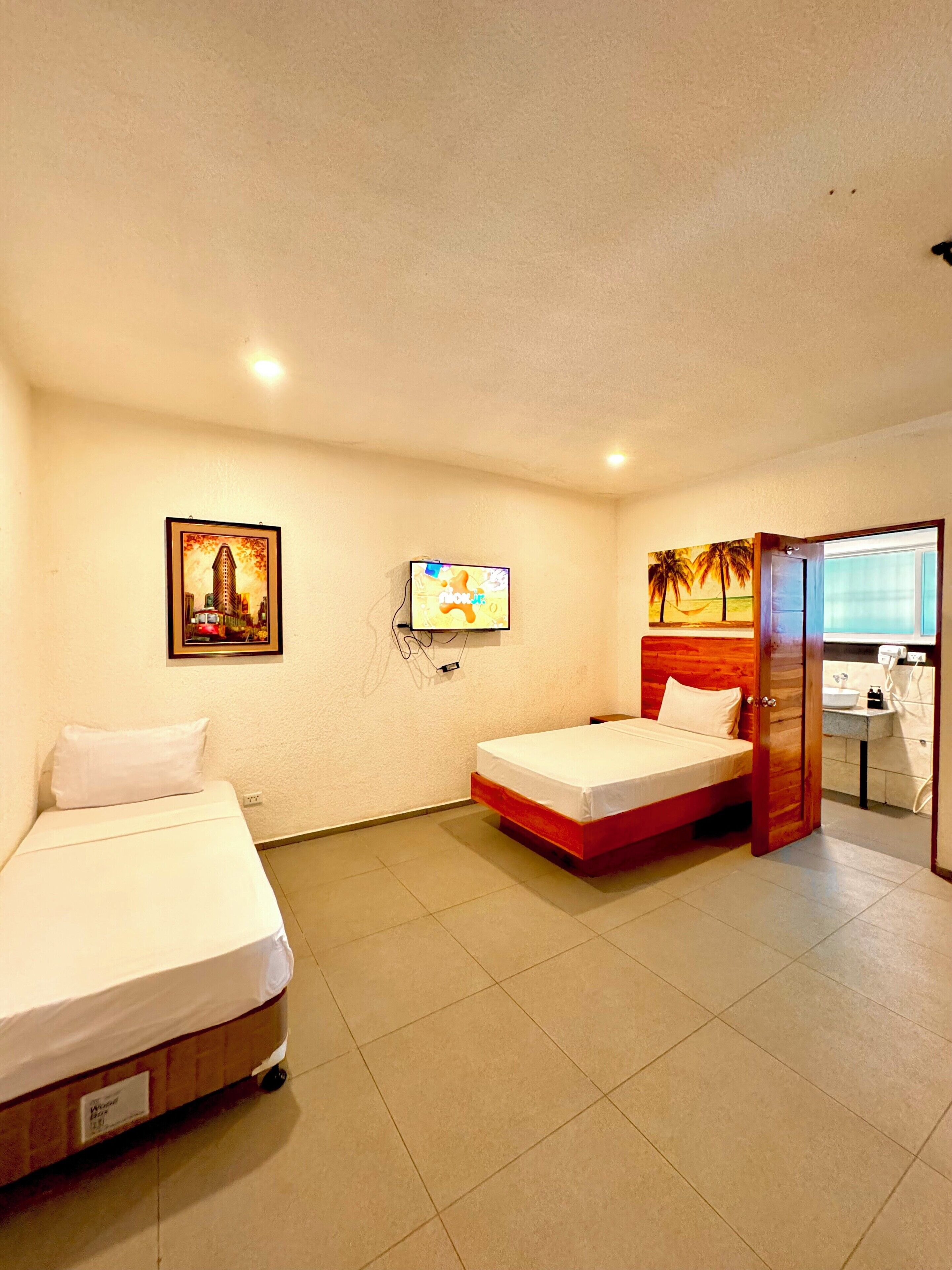 A hotel room with two single beds, a television, a painting, and a bathroom visible through an open door.