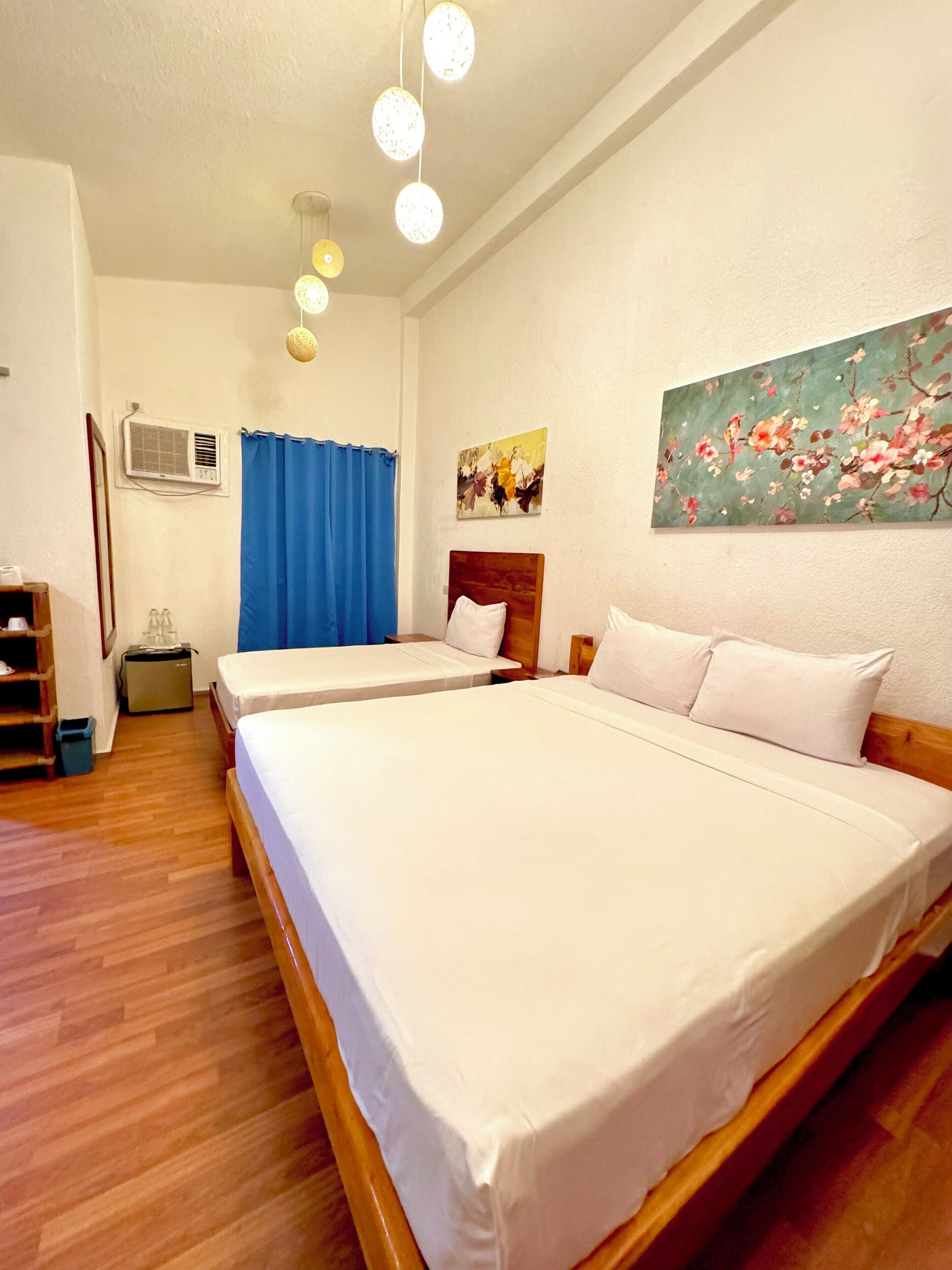 A hotel room with two beds, a blue curtain, a painting, and a ceiling light fixture.
