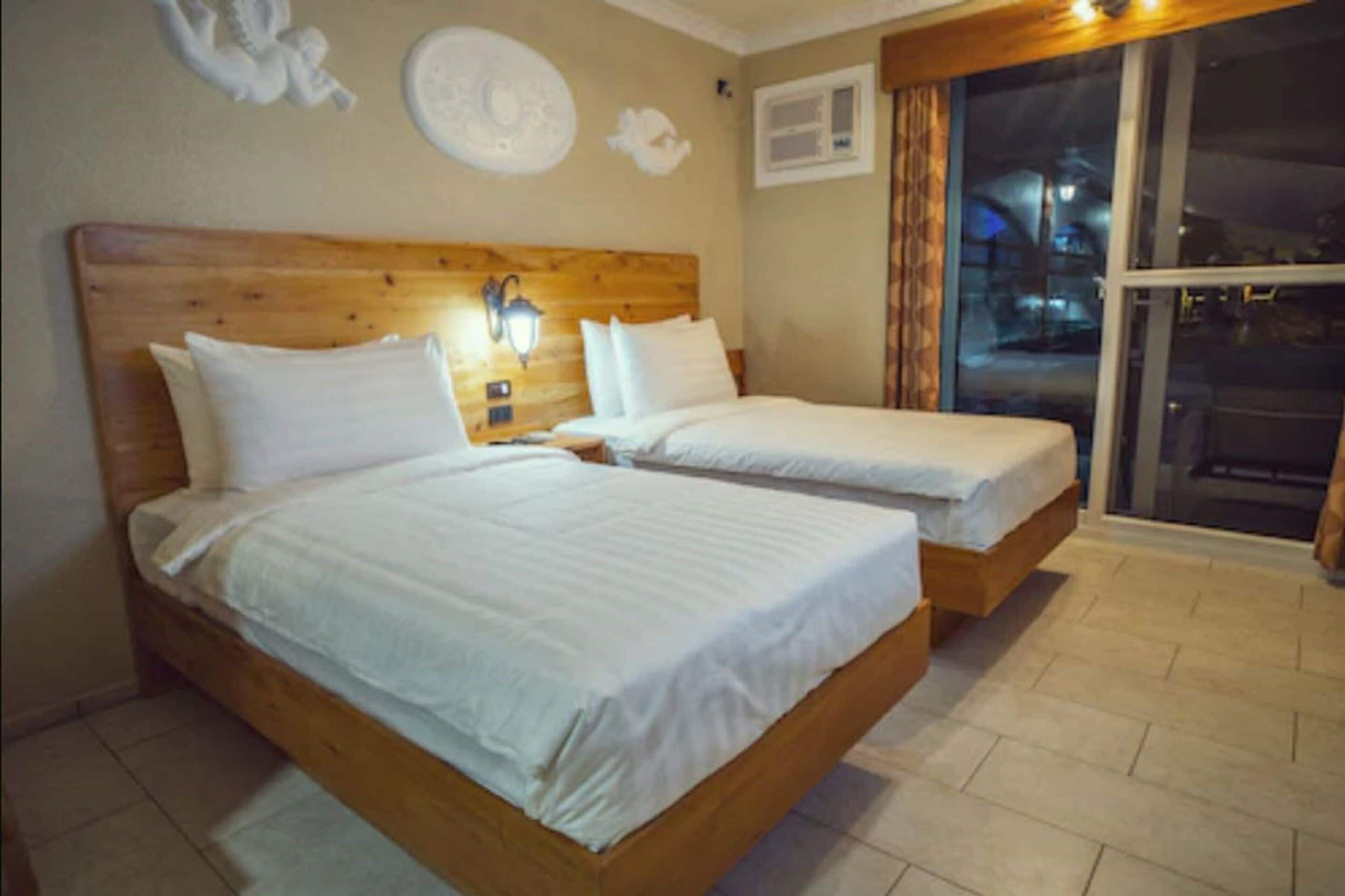 A room with two wooden beds, a nightstand with a lamp, and a window with a view of the outside.