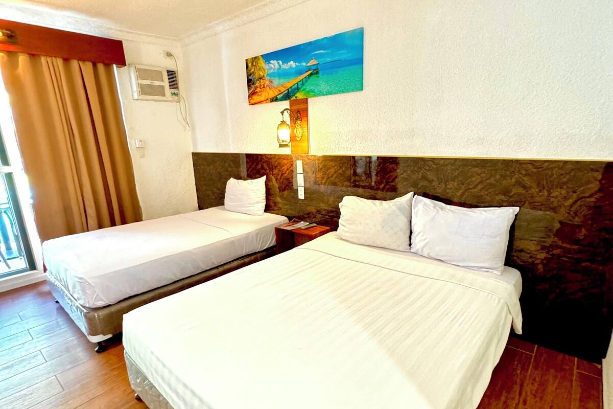 A hotel room with two beds, a painting of a beach scene, a wall-mounted air conditioner, and a window with curtains.